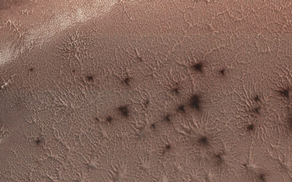 Chão do polo sul de Marte visto de cima, com marcas que lembram aranhas e manchas escuras.