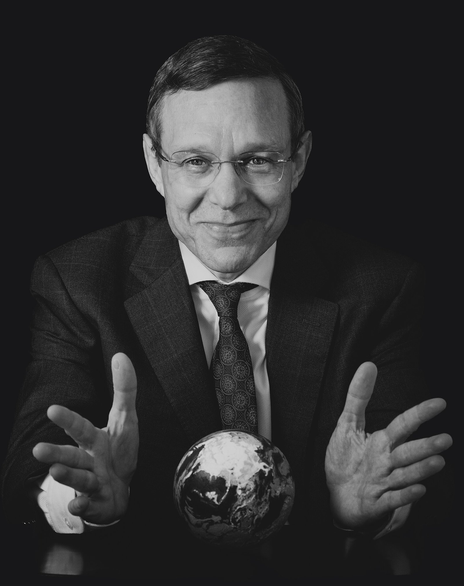 Avi Loeb, de óculos e terno, aparece com as mãos abertas ao redor de um globo decorativo, em retrato preto e branco.
