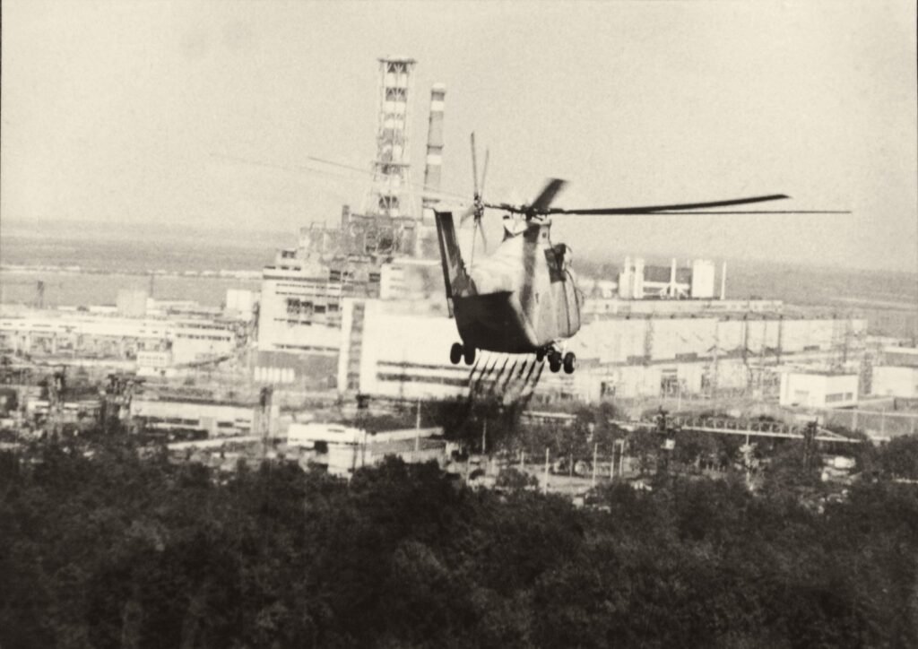 Um helicóptero voa baixo sobre árvores e solta um líquido; ao fundo, os prédios da usina e uma chaminé alta com armação.