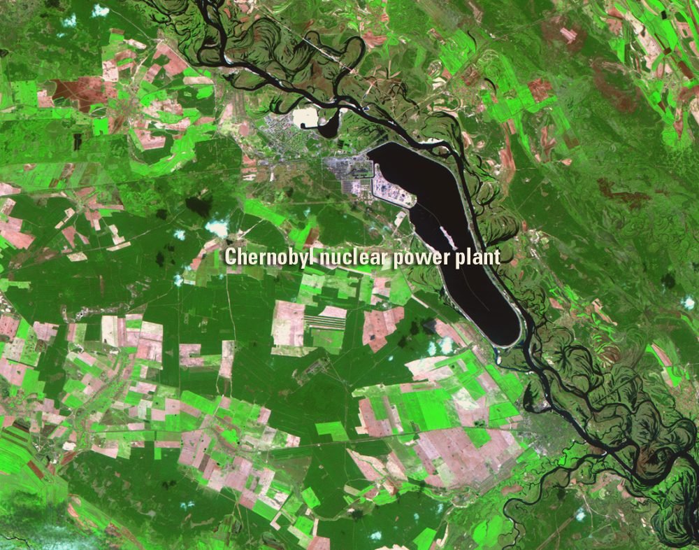 Imagem de satélite em cores falsas, com campos verdes e rosados; no centro, uma mancha preta comprida ao lado de um rio cheio de curvas; um rótulo em inglês indica a usina.