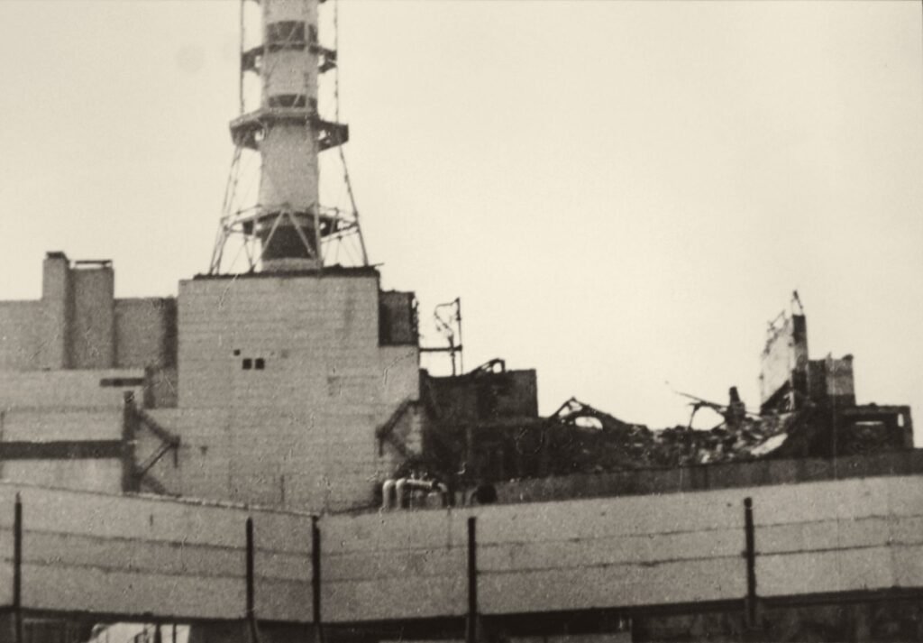 Um prédio industrial com a parte de cima arrebentada, ferros retorcidos à mostra, atrás de um muro; no alto, uma chaminé com armação de metal.