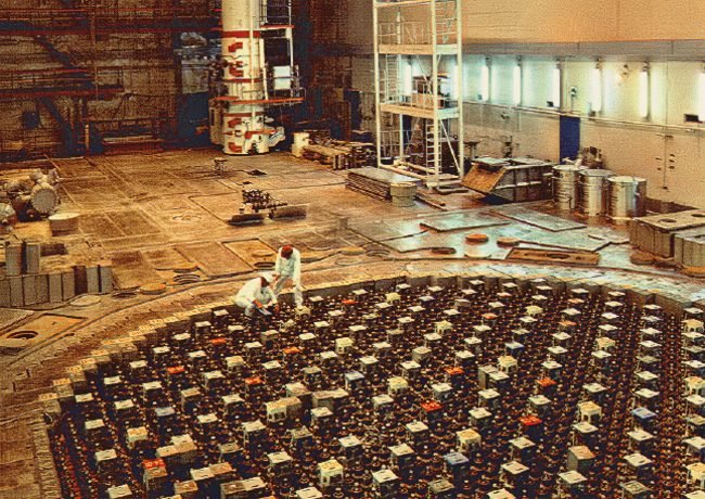 Um salão industrial enorme; o chão é um grande círculo formado por centenas de tampas quadradas; duas pessoas de roupa branca trabalham sobre ele.