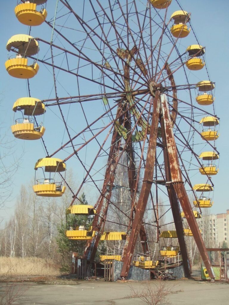 Uma roda-gigante enferrujada, com cabines amarelas, parada numa praça de concreto rachado; ao fundo, árvores sem folhas e um prédio.