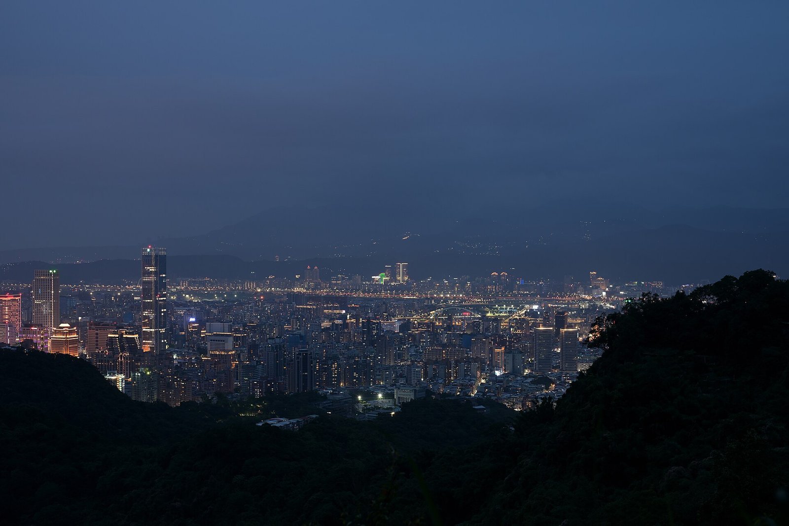 Panorama de Taipei ao entardecer, com prédios iluminados entre morros escuros e montanhas no horizonte.