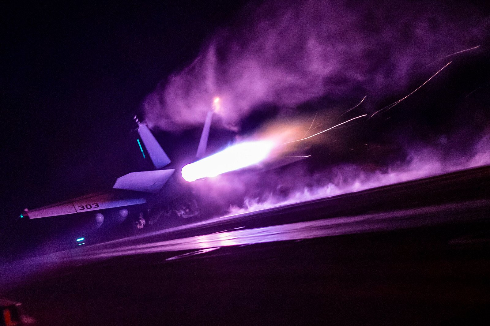 Fotografia noturna de um F/A-18E em decolagem no USS Nimitz, com chama do motor e vapor iluminado sobre o convés.