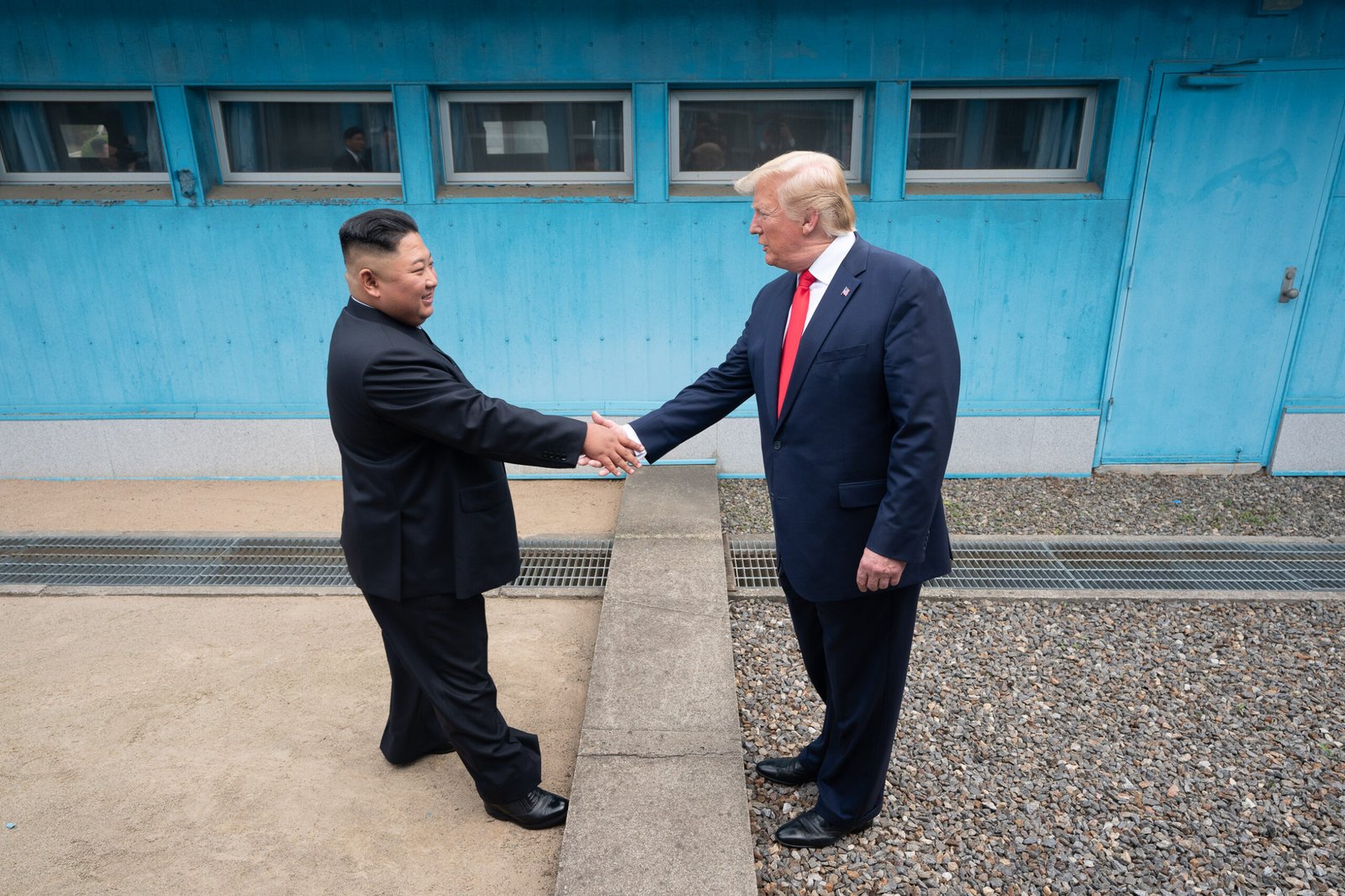 Donald Trump e Kim Jong-un apertam as mãos na linha de demarcação, com edifícios de Panmunjom ao fundo.