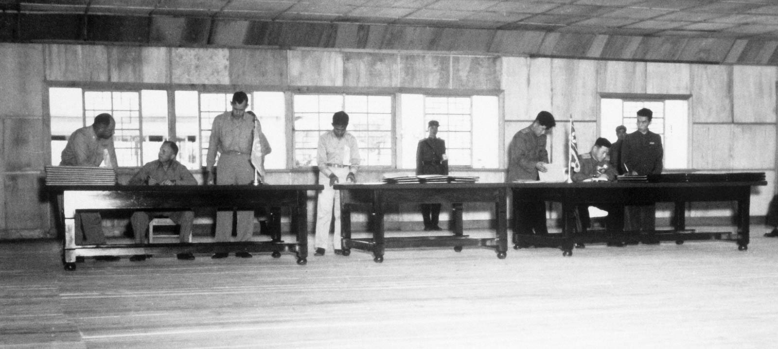 Fotografia em preto e branco dos delegados militares William Harrison e Nam Il sentados à mesa durante a assinatura do armistício.