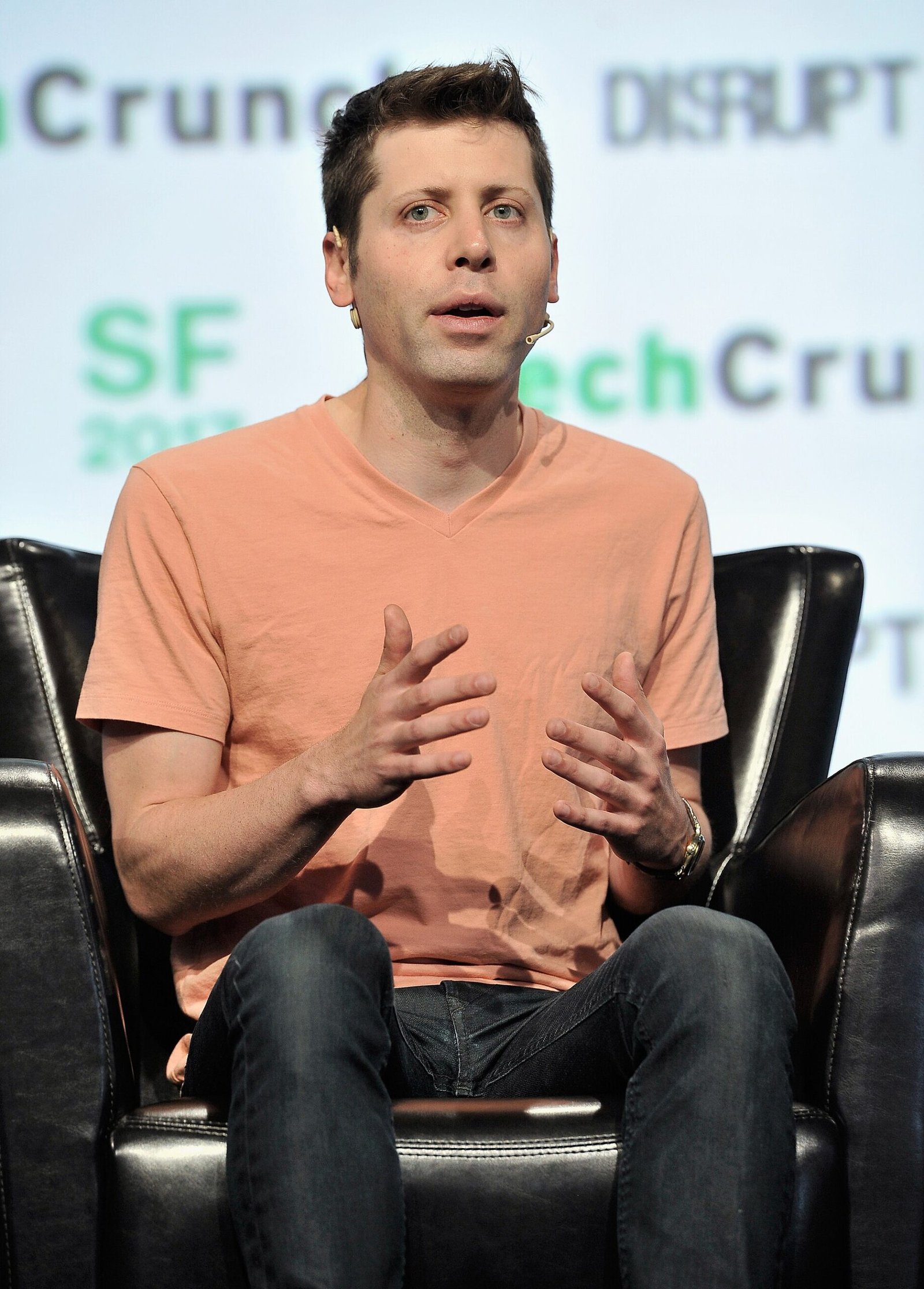 Sam Altman sentado e gesticulando com as mãos, usando camiseta laranja e microfone junto ao rosto, com painel verde ao fundo.