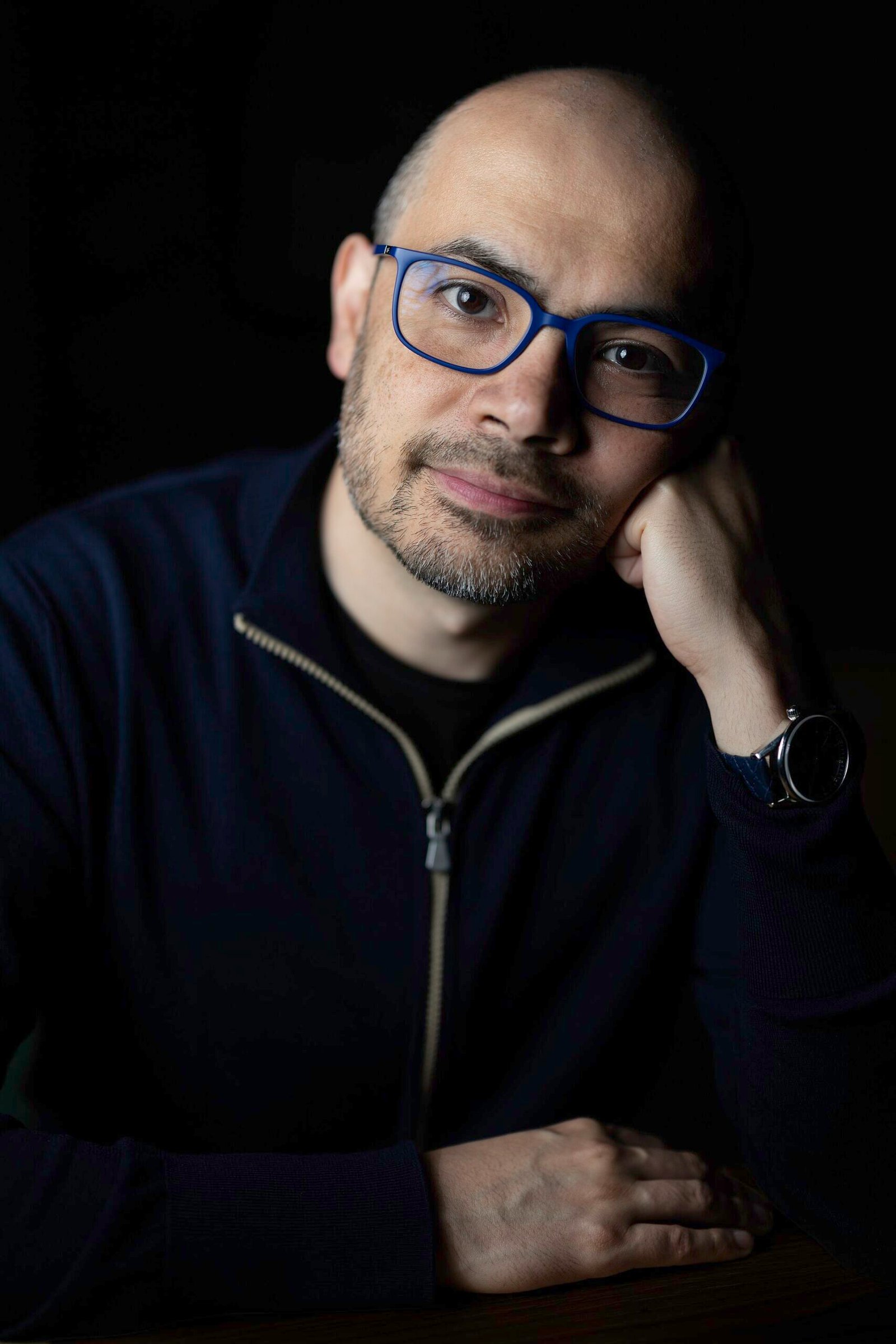 Retrato de Demis Hassabis usando óculos, com fundo escuro.