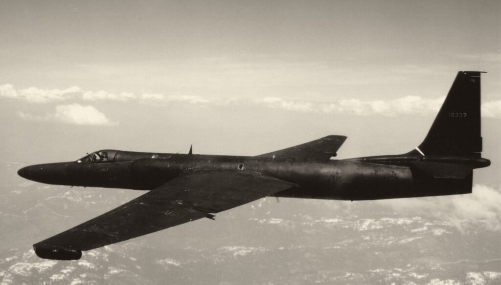 Foto em preto e branco: um avião escuro, de asas muito compridas e finas, voa acima das nuvens.