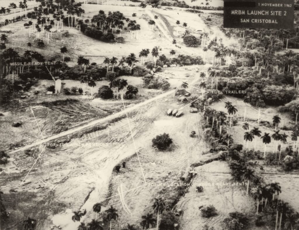Foto aérea em preto e branco: um terreno de terra revirada, com estradas, palmeiras e poucos veículos; etiquetas em inglês apontam lugares vazios.