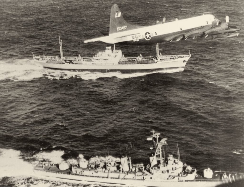 Foto aérea em preto e branco: um avião branco de quatro motores voa baixo sobre um navio cargueiro; em primeiro plano, um navio de guerra.