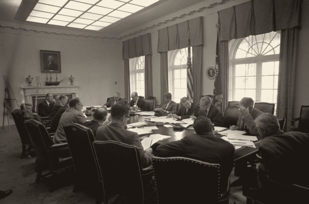 Foto em preto e branco: mais de dez homens de terno sentados em volta de uma mesa comprida, numa sala de janelas altas com cortinas.