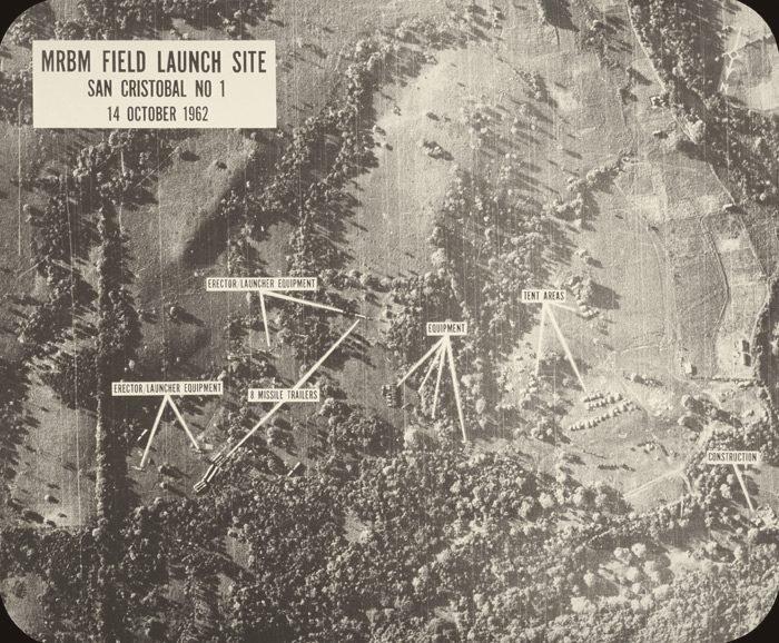 Foto aérea em preto e branco de um terreno com árvores e clareiras; etiquetas em inglês, com setas, apontam pequenos objetos no chão.