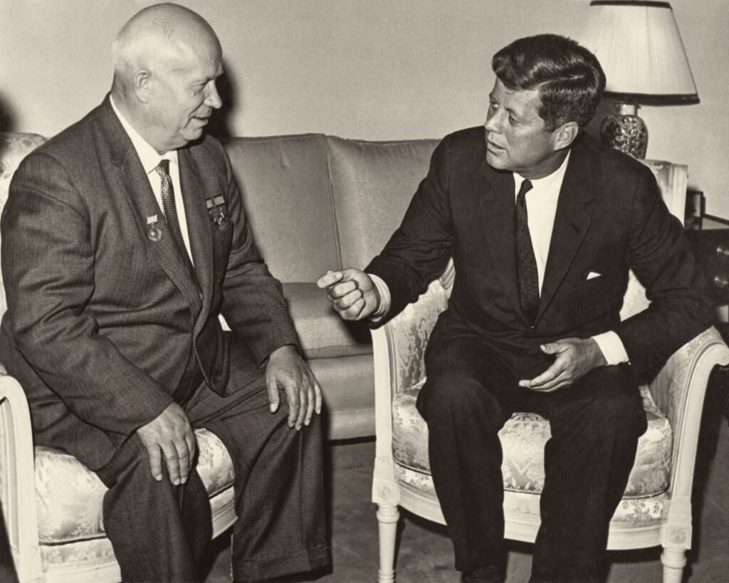 Foto em preto e branco: dois homens de terno conversam sentados em poltronas claras; o da esquerda, calvo, sorri; o da direita, mais jovem, olha para ele.