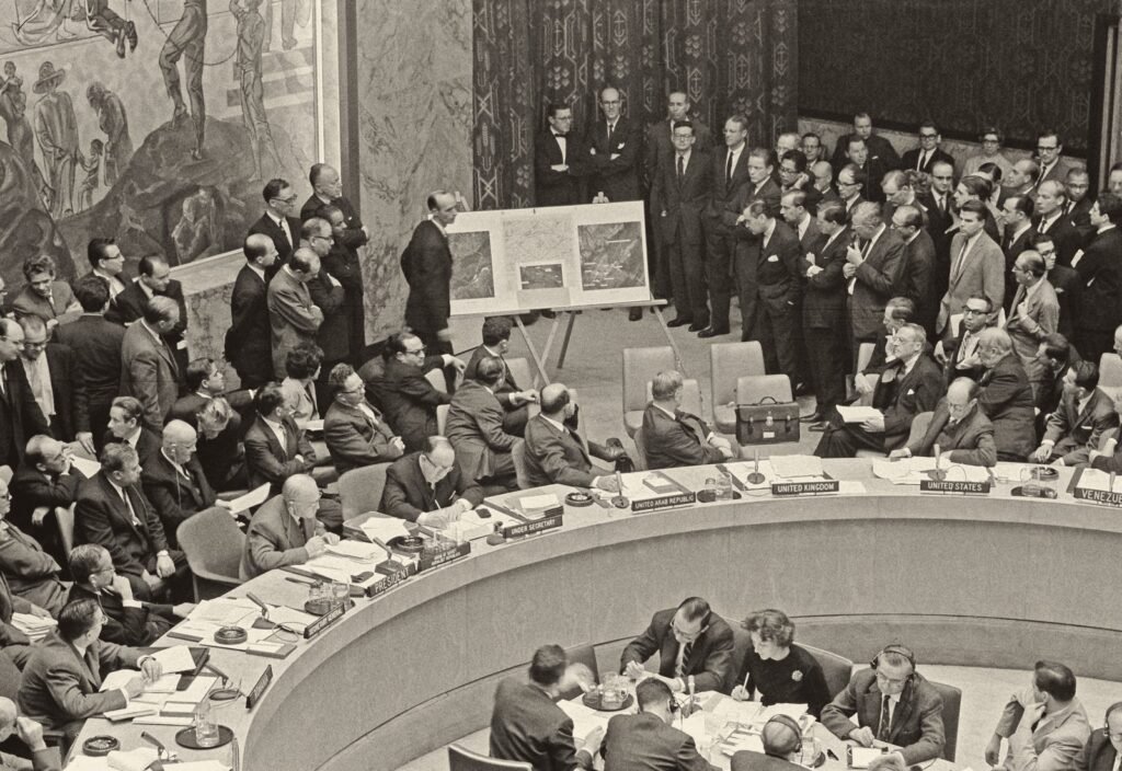 Foto em preto e branco: sala lotada, com uma grande mesa em curva; ao fundo, num cavalete, três painéis com fotos aéreas, e um homem de pé ao lado.