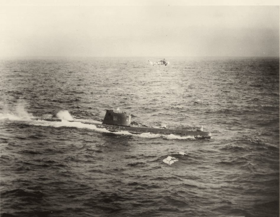 Foto em preto e branco, tirada do alto: um submarino navega na superfície de um mar agitado; acima dele, ao longe, um helicóptero.