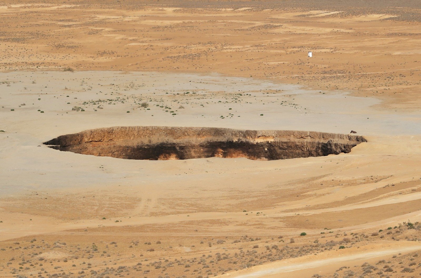 Uma cavidade oval interrompe o terreno arenoso, com dunas e vegetação baixa ao redor.