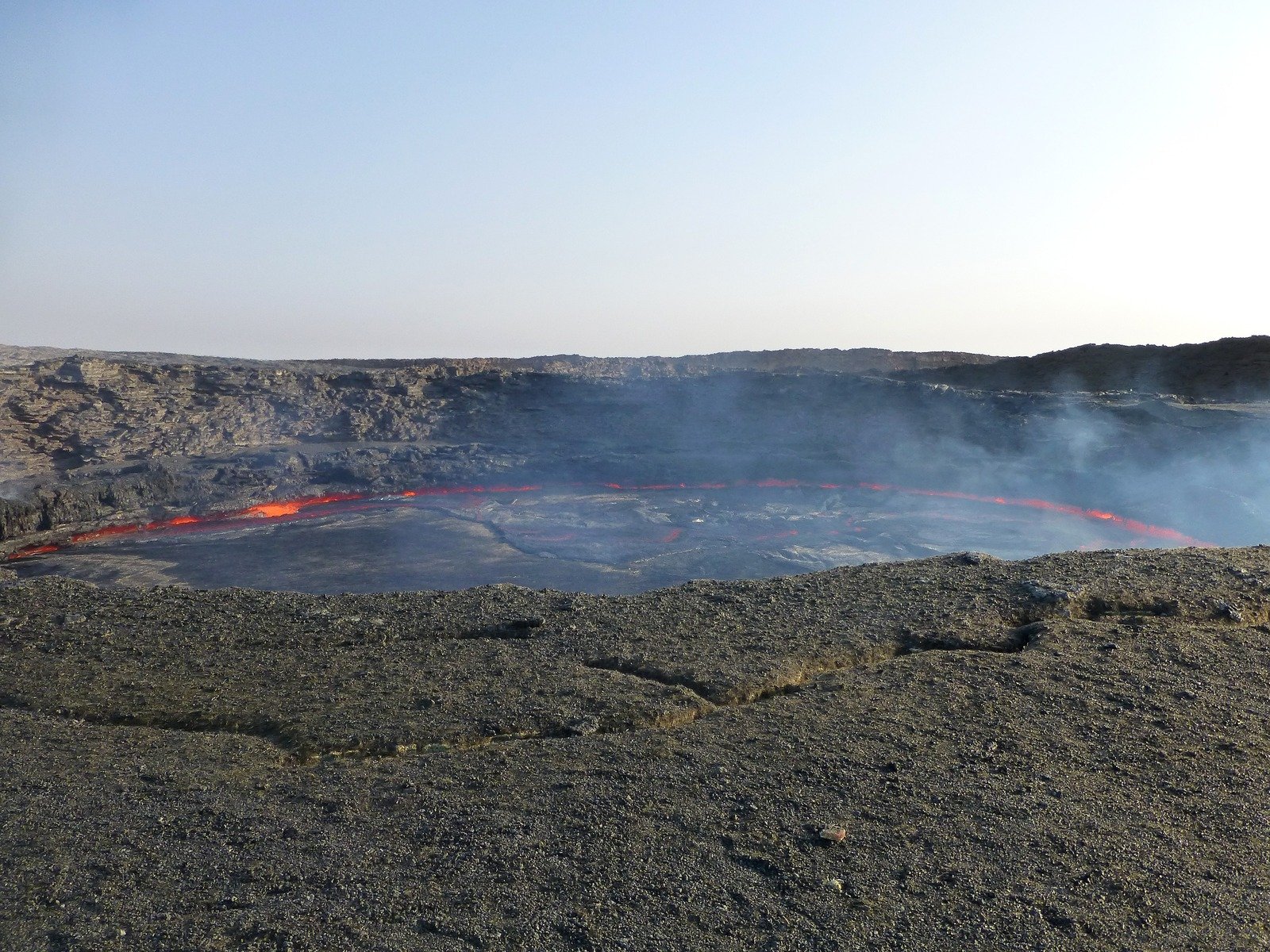 Uma cratera larga contém crosta escura, lava vermelha nas bordas e uma névoa de gases acima.
