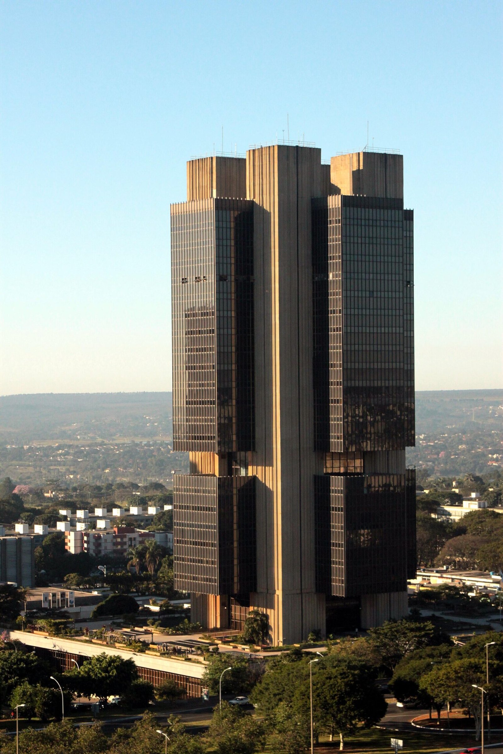 Edifício alto da sede do Banco Central em Brasília, com vegetação e outros prédios ao redor.