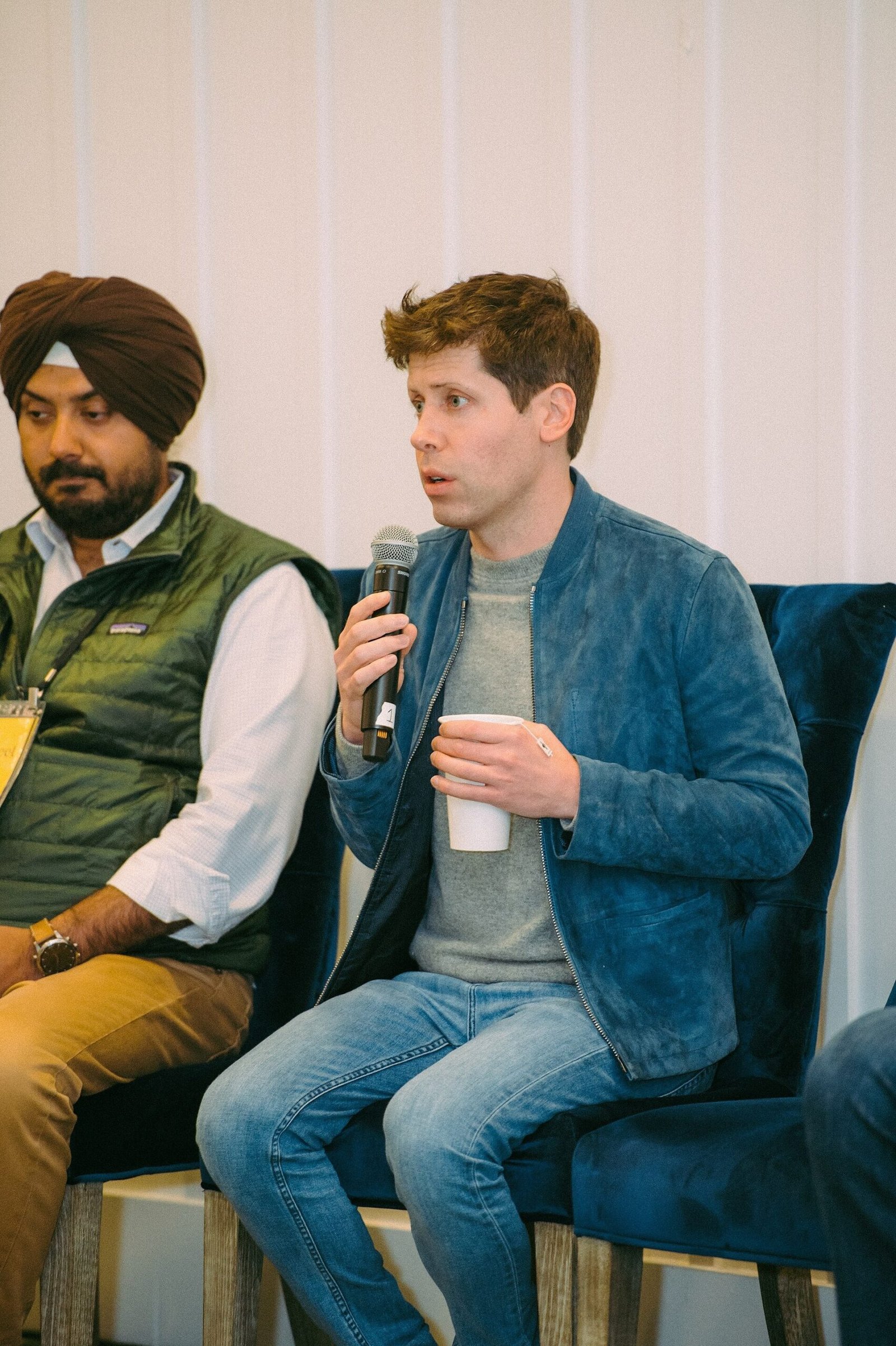 Sam Altman segura um microfone sentado ao lado de outro participante de um evento.