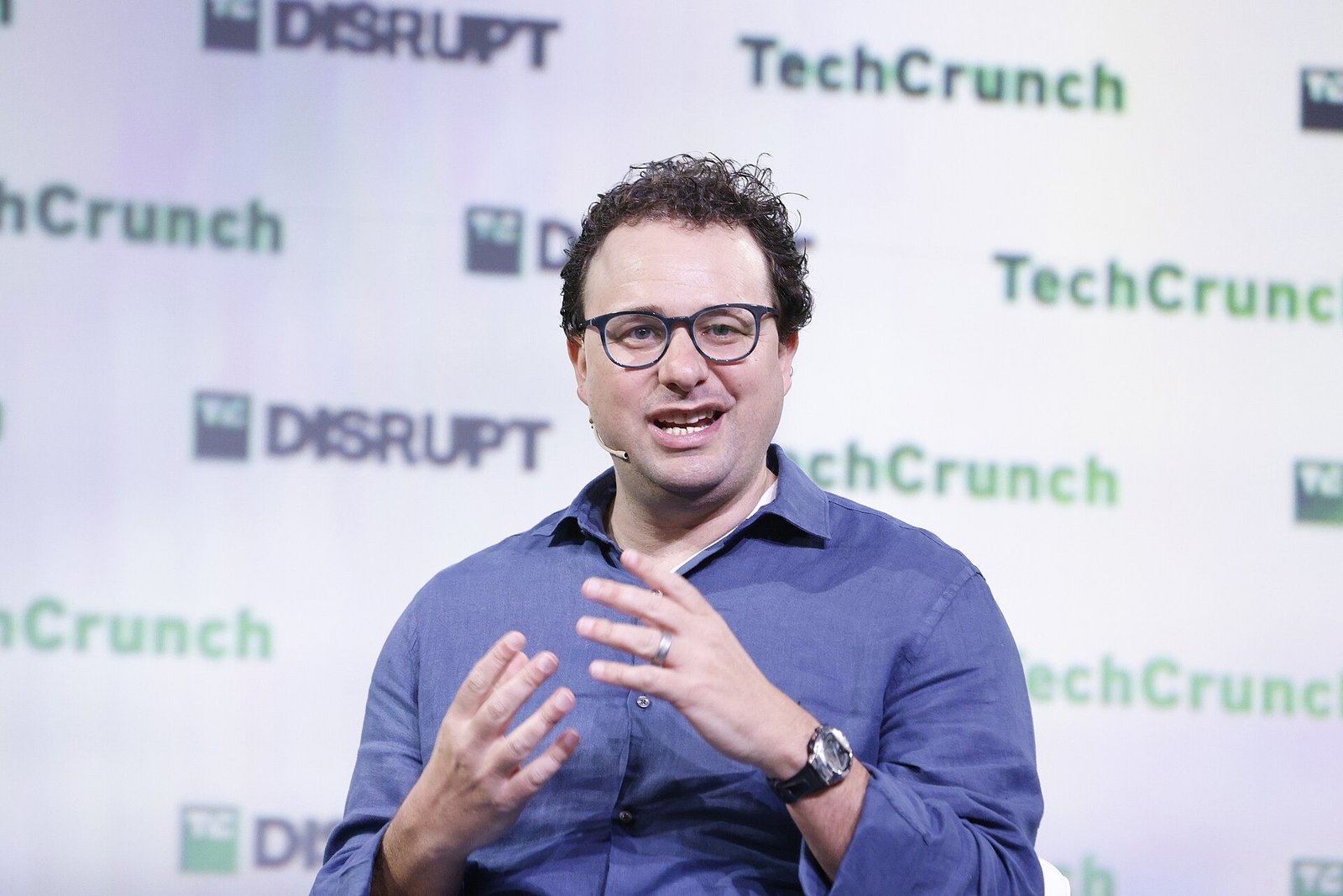 Dario Amodei, com óculos e camisa azul, fala sentado diante de um painel da TechCrunch.