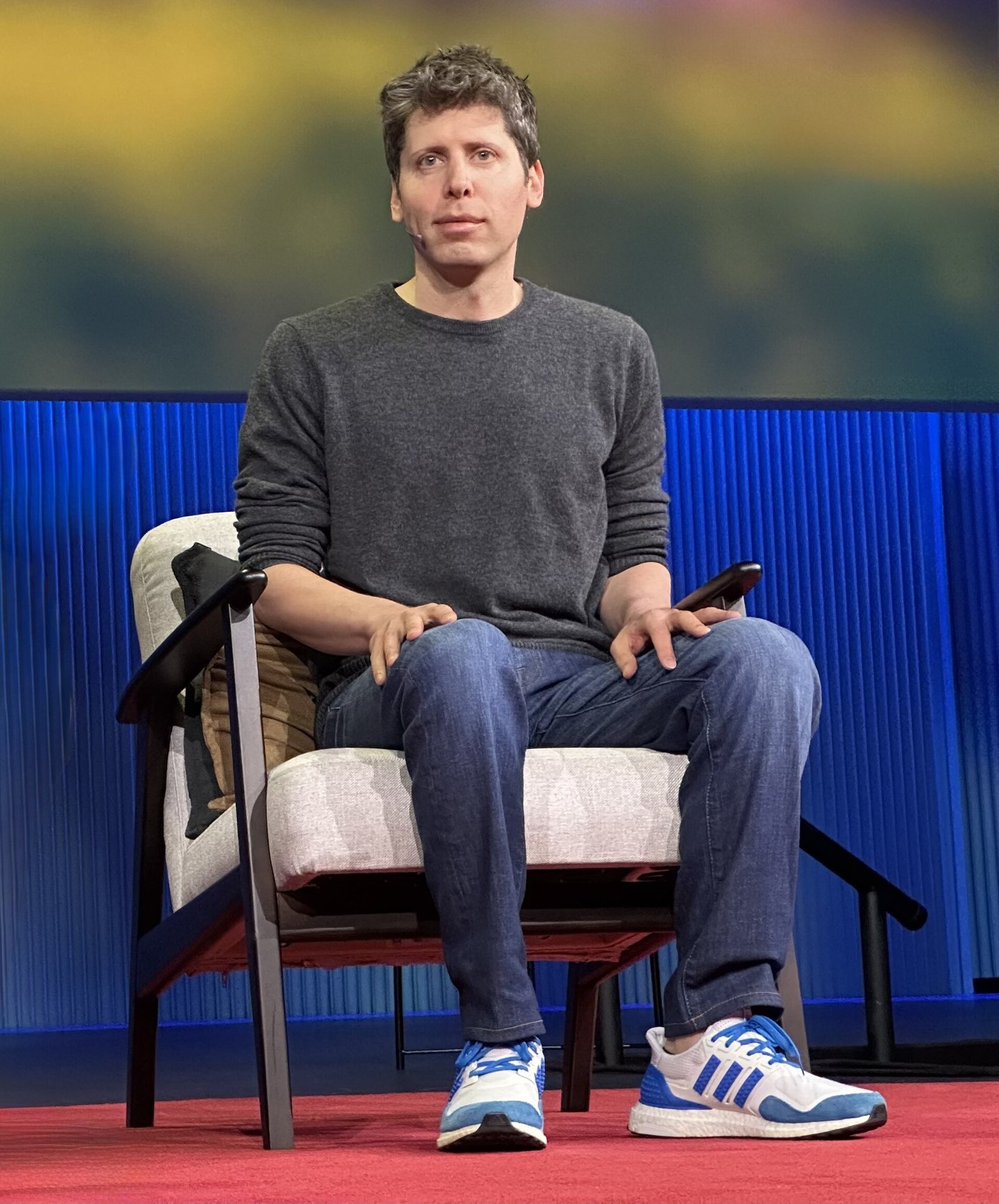 Sam Altman, com suéter cinza, está sentado em uma poltrona no palco, segurando um telefone.