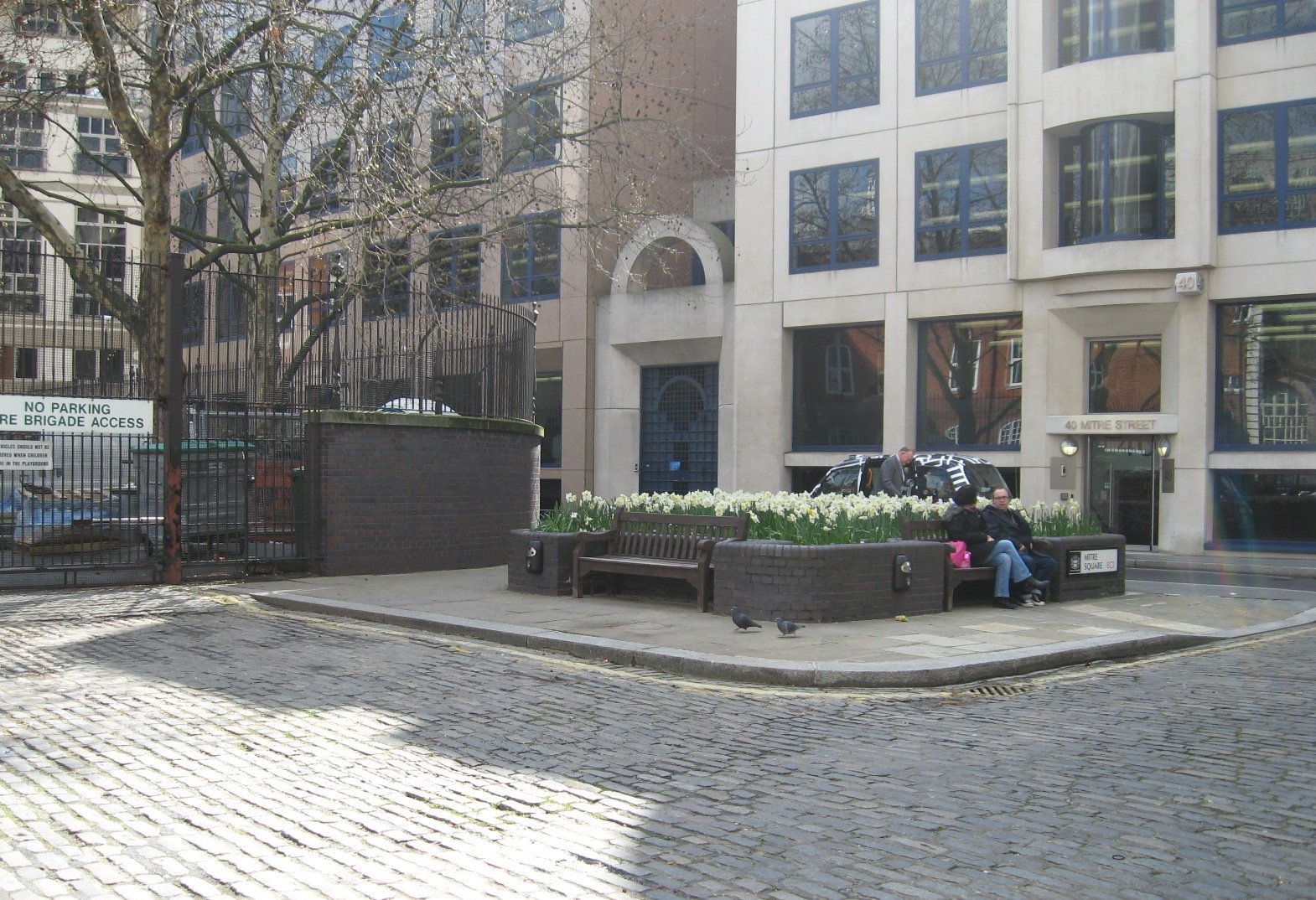 Trecho de Mitre Square com bancos, canteiros, árvores e fachadas de escritórios modernos, fotografado de dia.