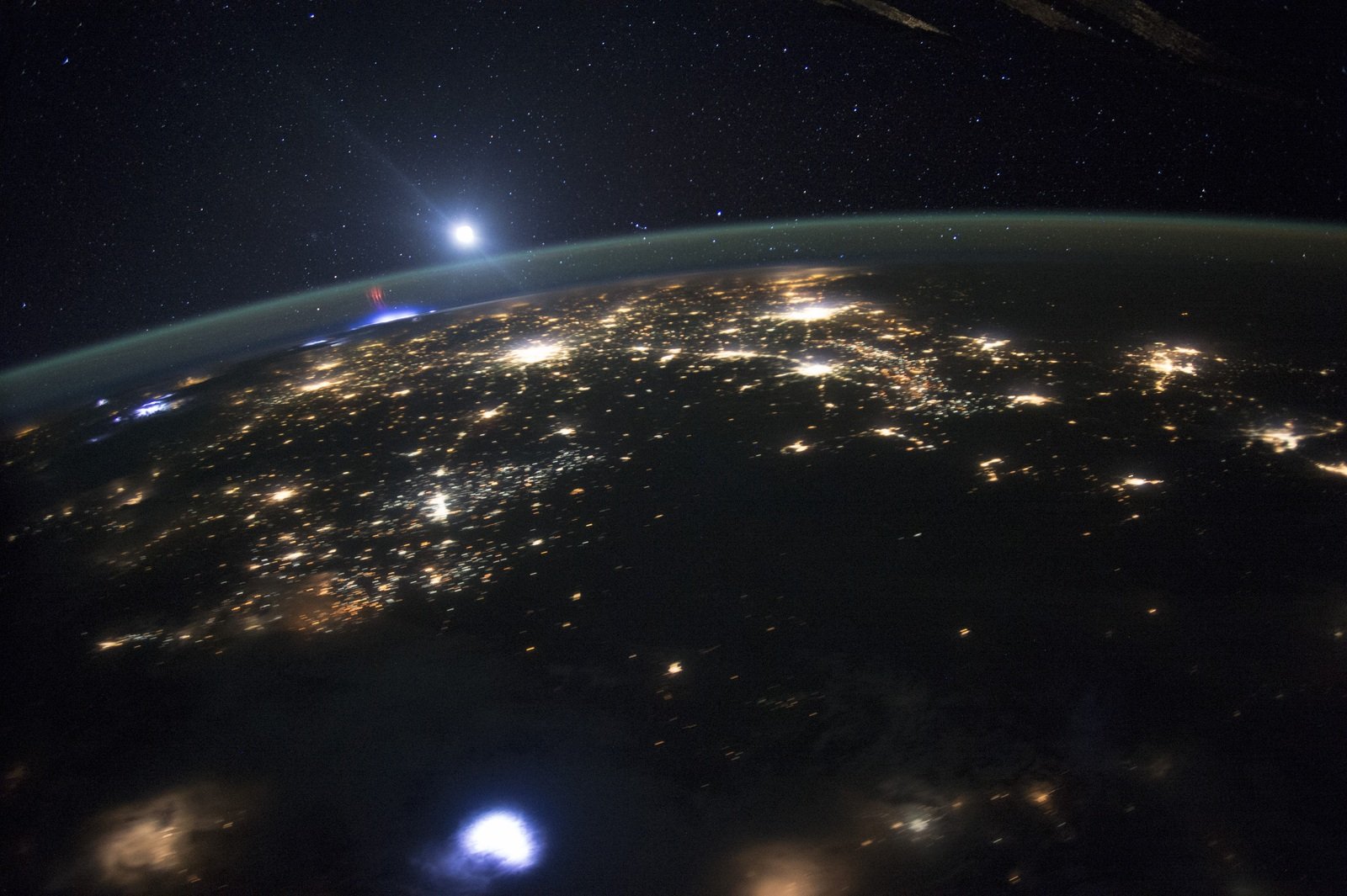 A borda curva da Terra aparece entre estrelas e luzes de cidades; perto da Lua, pequenos filamentos vermelhos surgem acima de uma tempestade azulada.