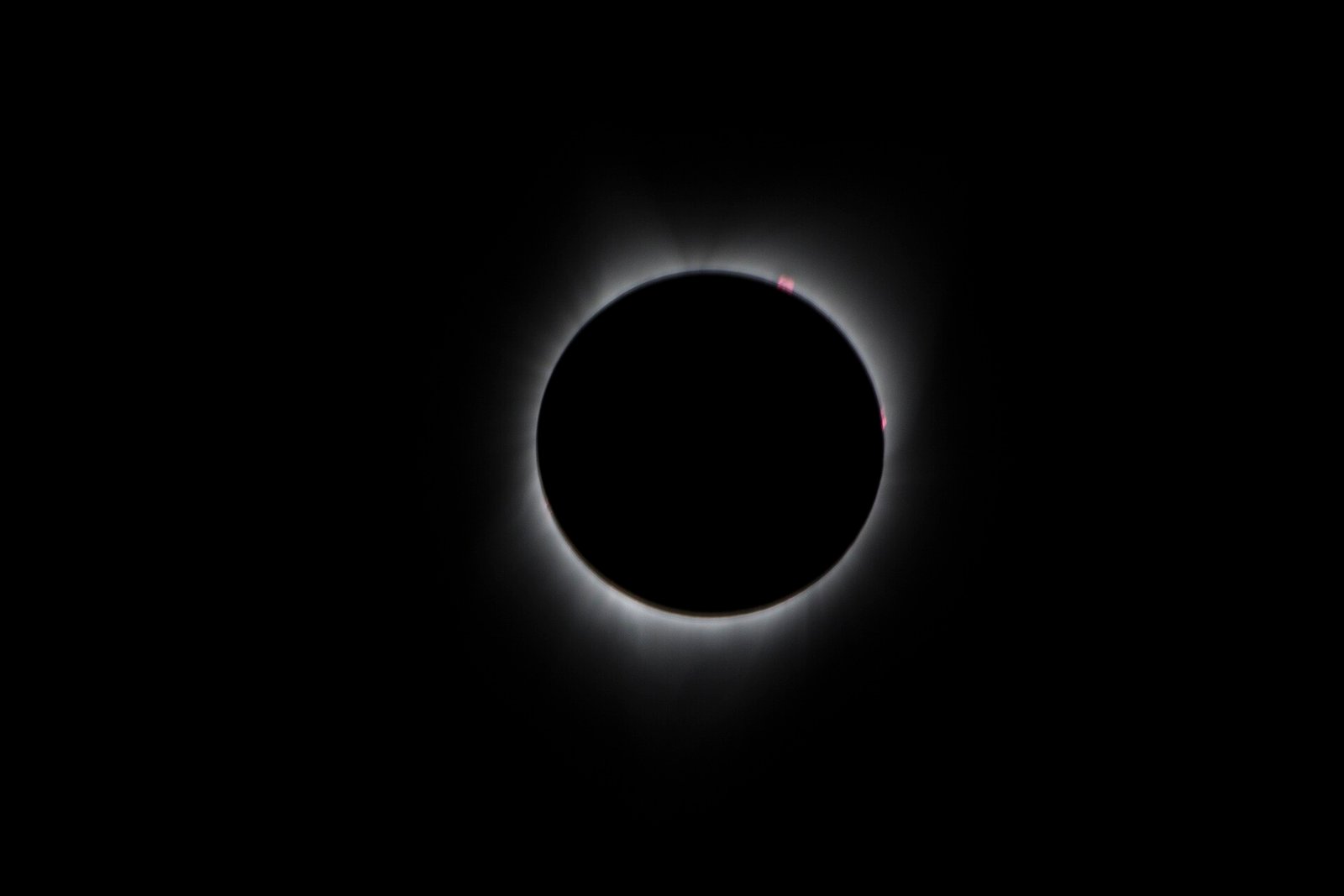 Um disco negro recobre o Sol, cercado por uma coroa branca de luz; pequenas estruturas avermelhadas aparecem junto à borda.