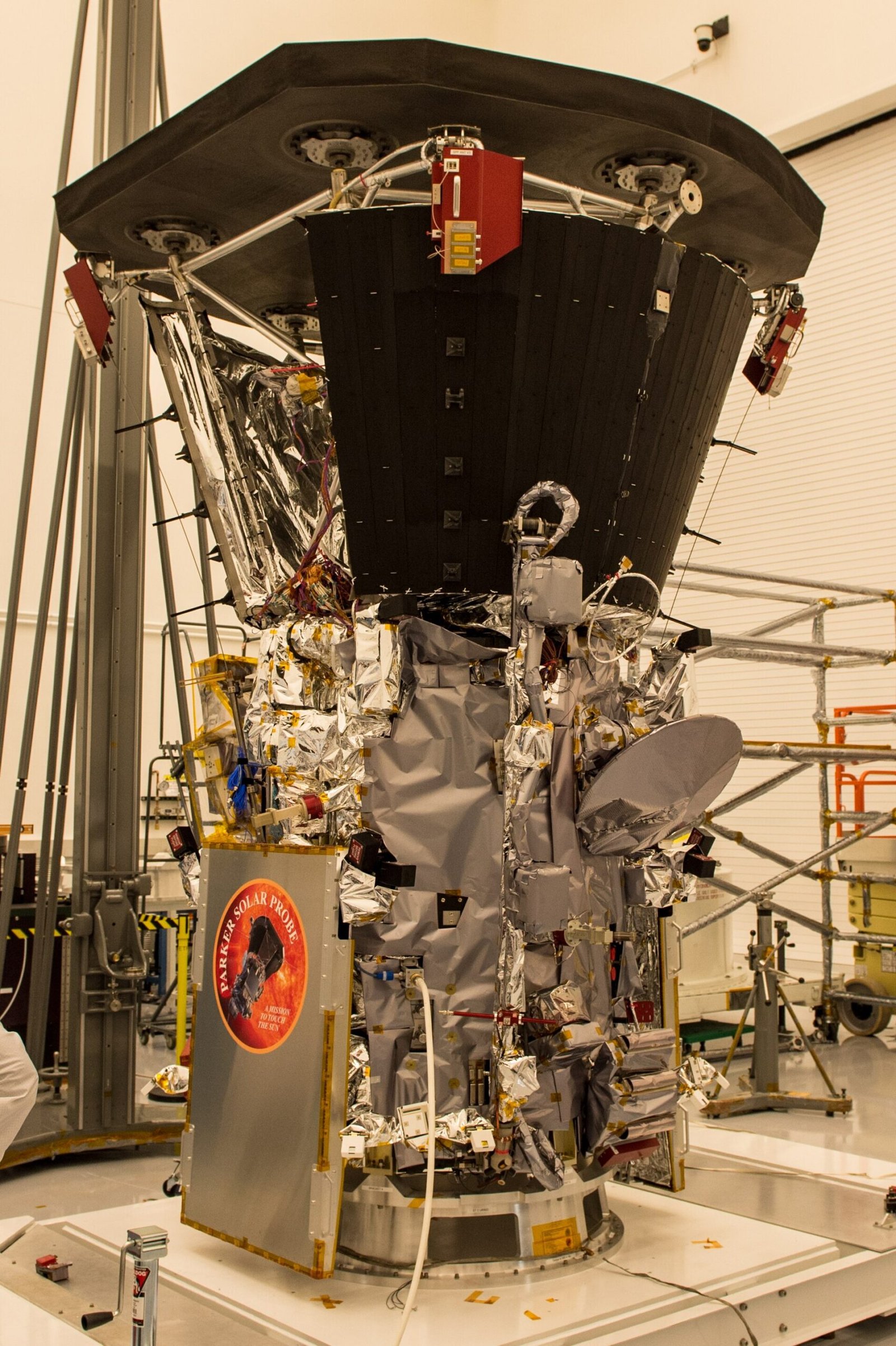 A Parker Solar Probe aparece numa sala limpa, com um escudo escuro na parte superior, estruturas metálicas e painéis ao redor.
