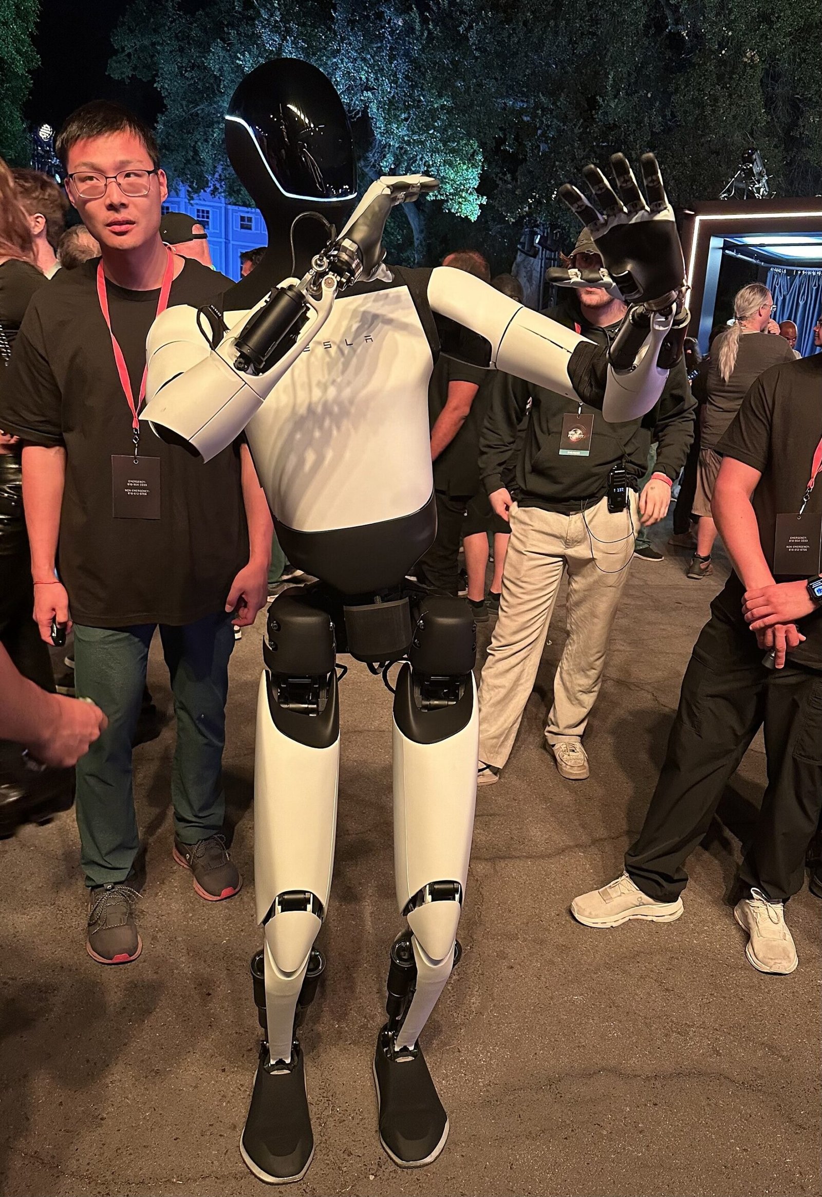 Robô humanoide Tesla Optimus branco e preto, com as mãos levantadas, entre participantes de um evento.