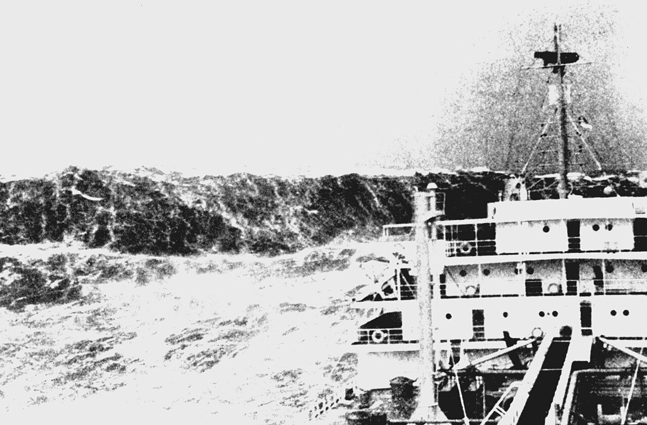 Fotografia em preto e branco de um navio enfrentando uma grande onda, com o convés e a superestrutura à direita.
