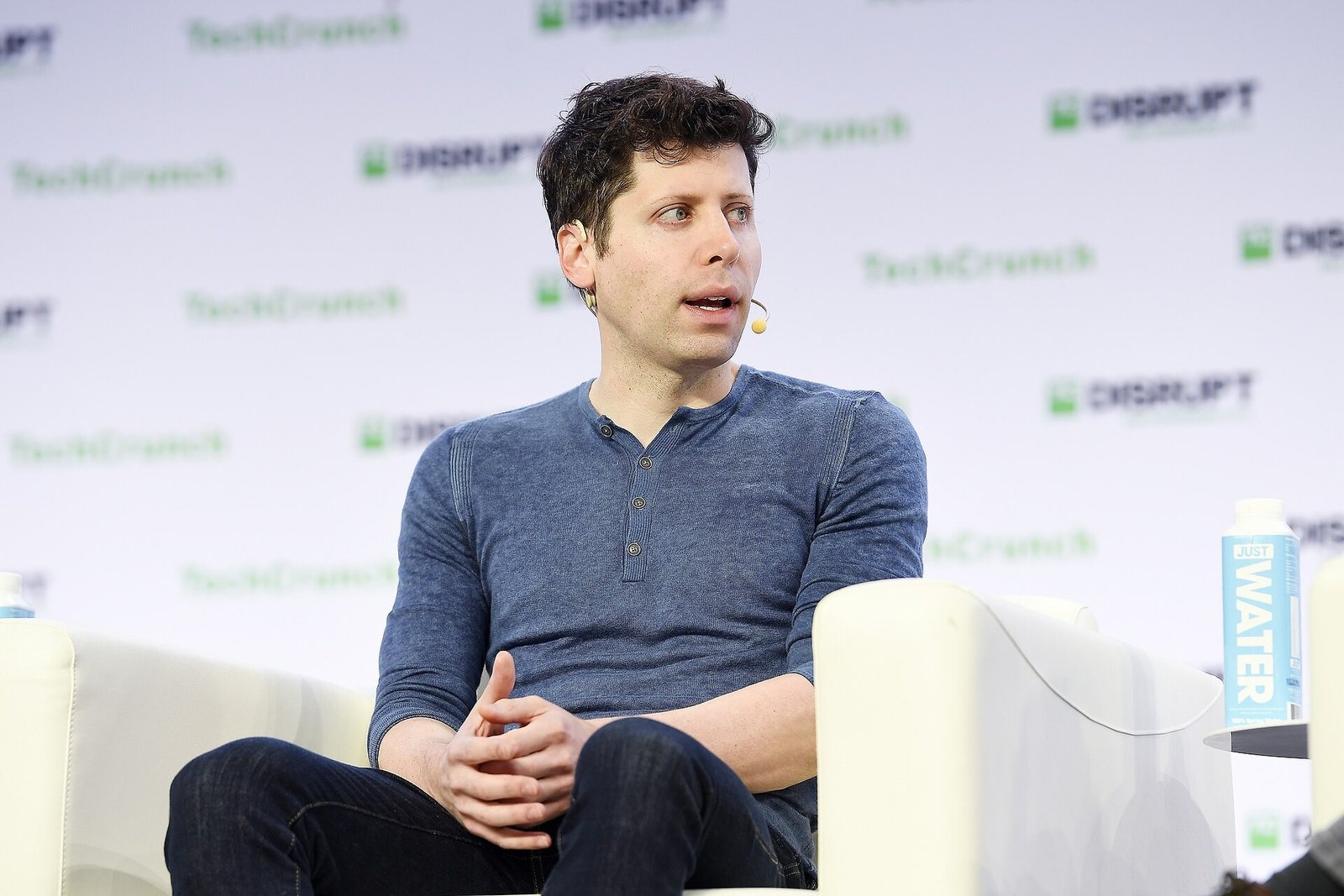 Sam Altman, com camisa azul e microfone de rosto, está sentado numa poltrona diante do painel da TechCrunch.