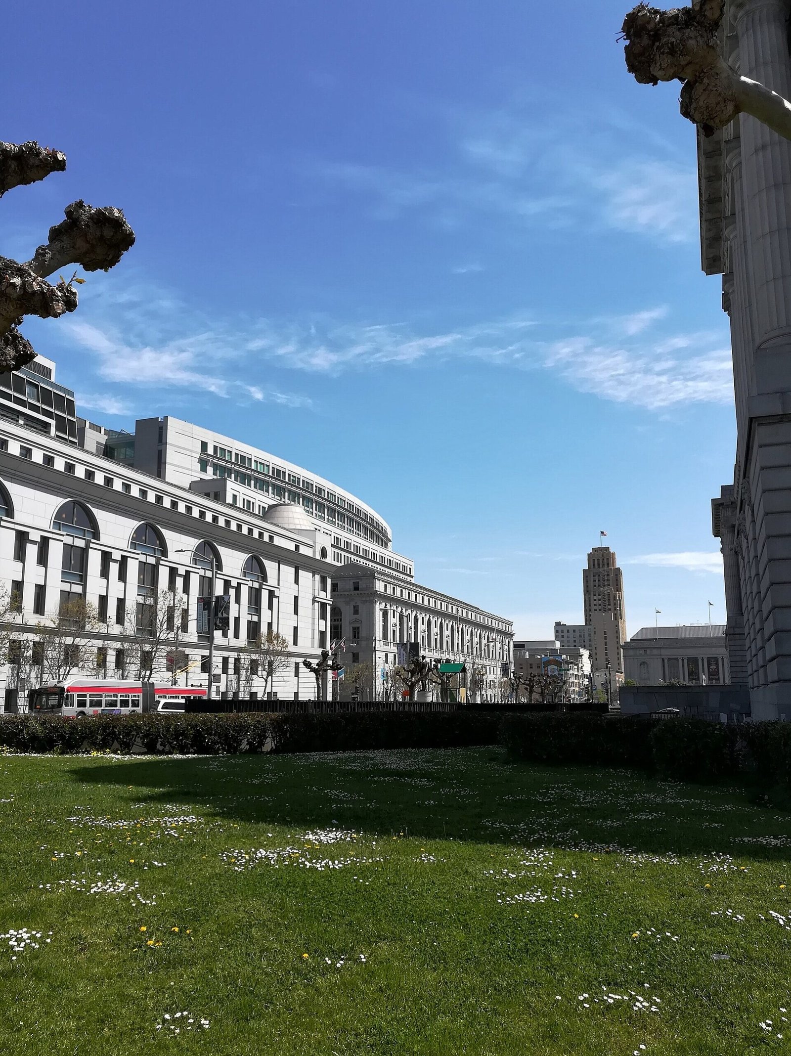 Edifícios do Civic Center de San Francisco aparecem atrás de um gramado e de uma rua por onde passa um bonde.