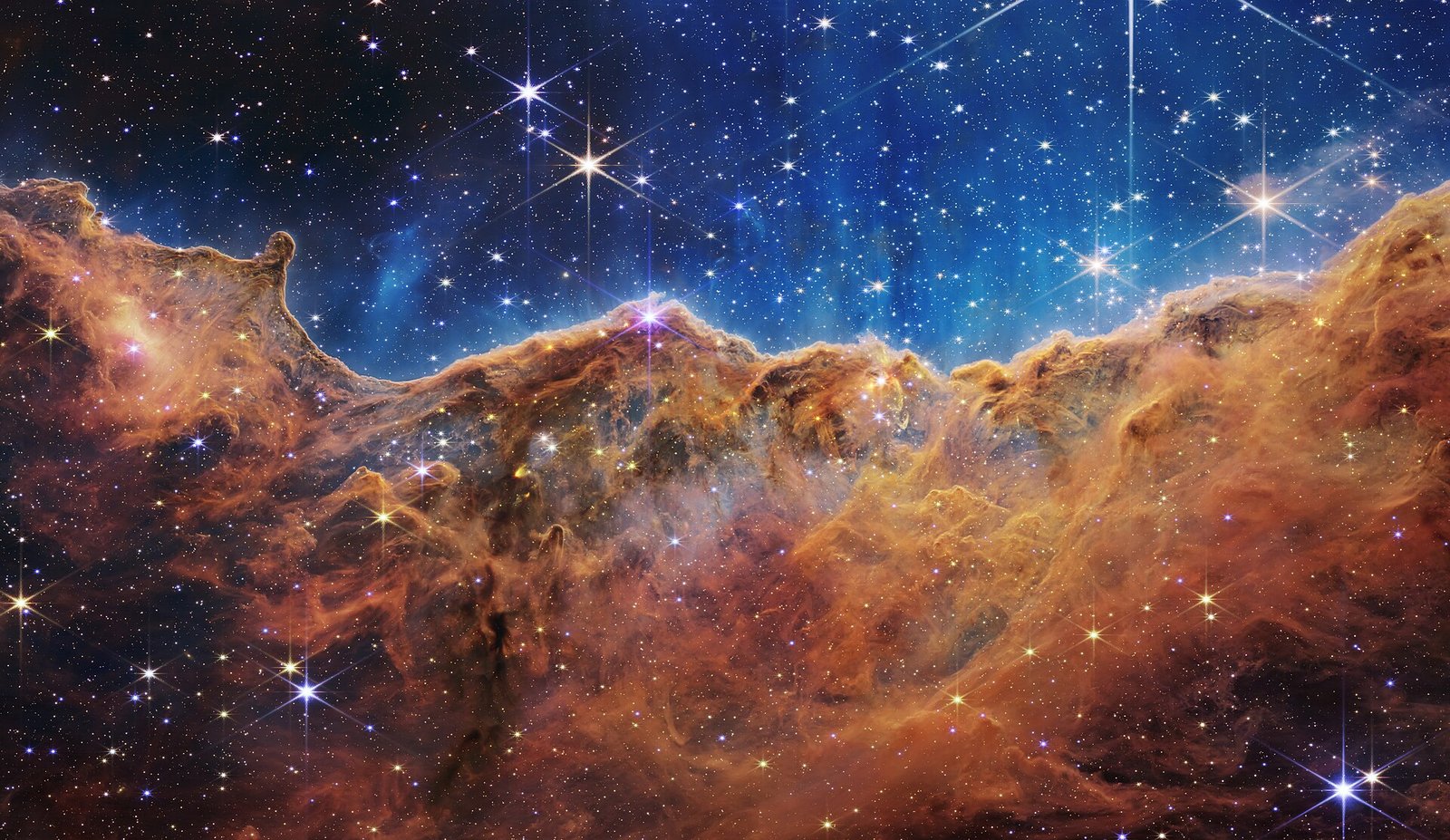 Uma parede ondulada de gás e poeira alaranjada ocupa a parte inferior da cena, sob um campo azul repleto de estrelas.