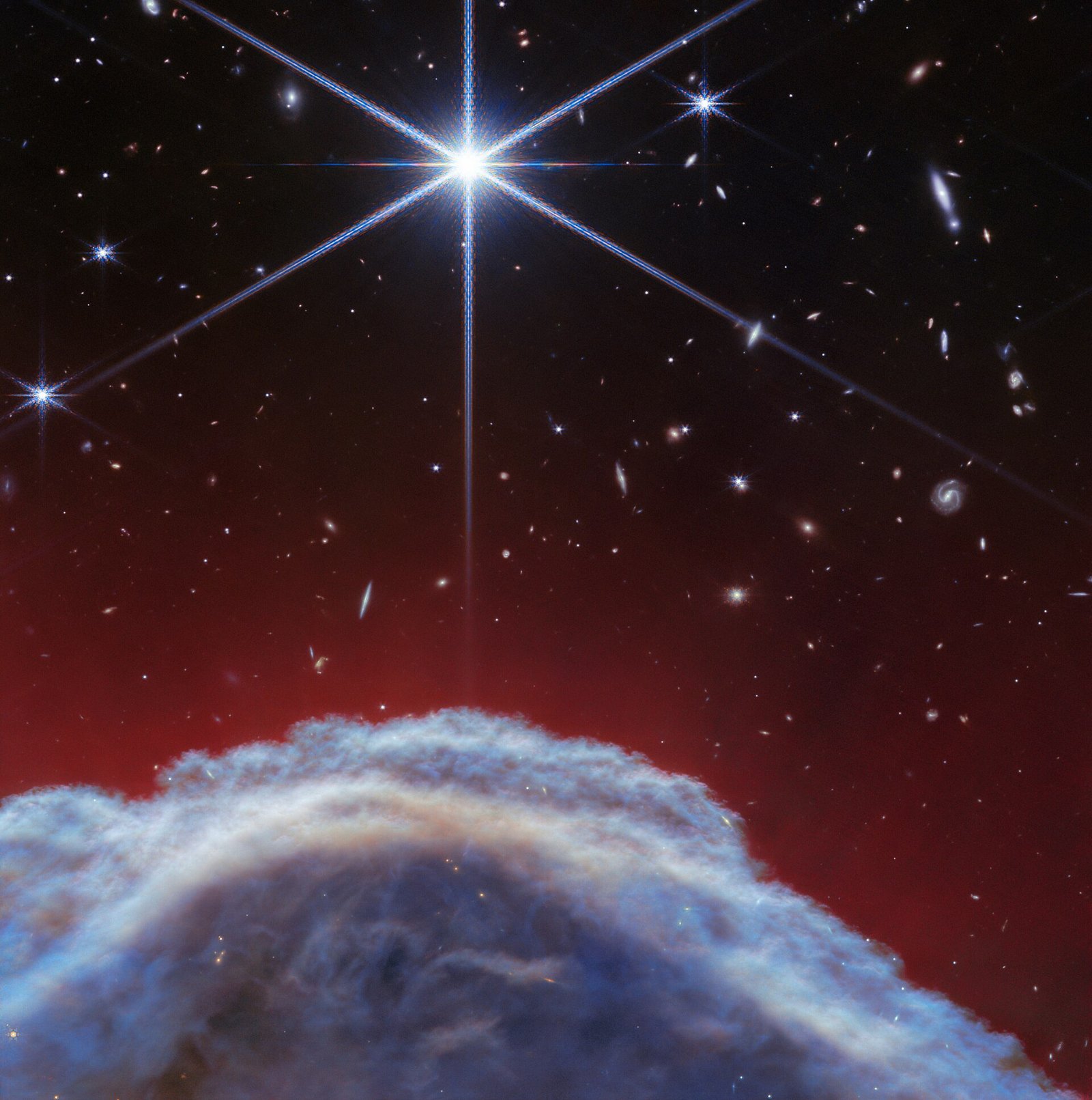 A borda cinza-azulada de uma nuvem curva aparece sob filamentos vermelhos e estrelas; uma estrela brilhante com longas espículas está no alto.