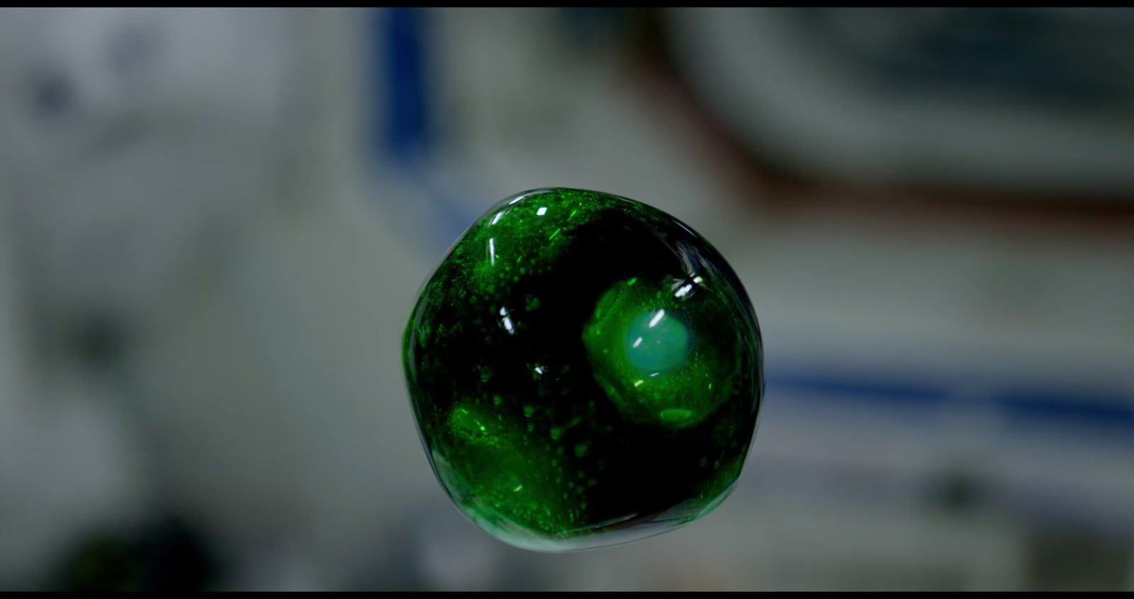 Uma esfera de água verde, cheia de pequenas bolhas, flutua diante dos equipamentos desfocados da Estação Espacial.