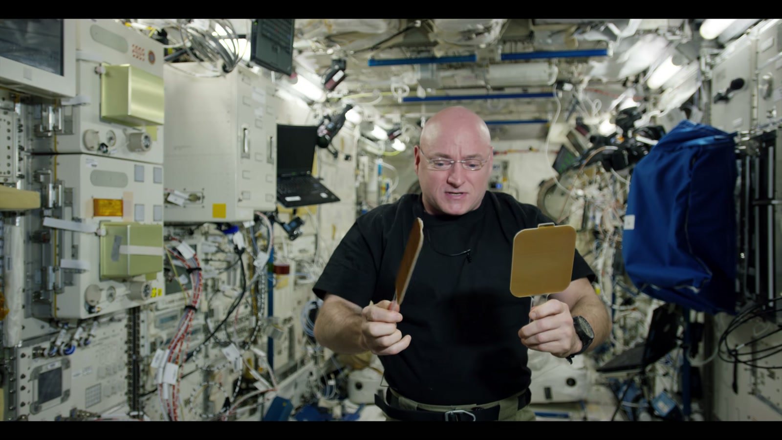 Scott Kelly, de camiseta preta e óculos, segura duas pequenas raquetes diante do corpo, entre cabos e equipamentos da Estação Espacial.