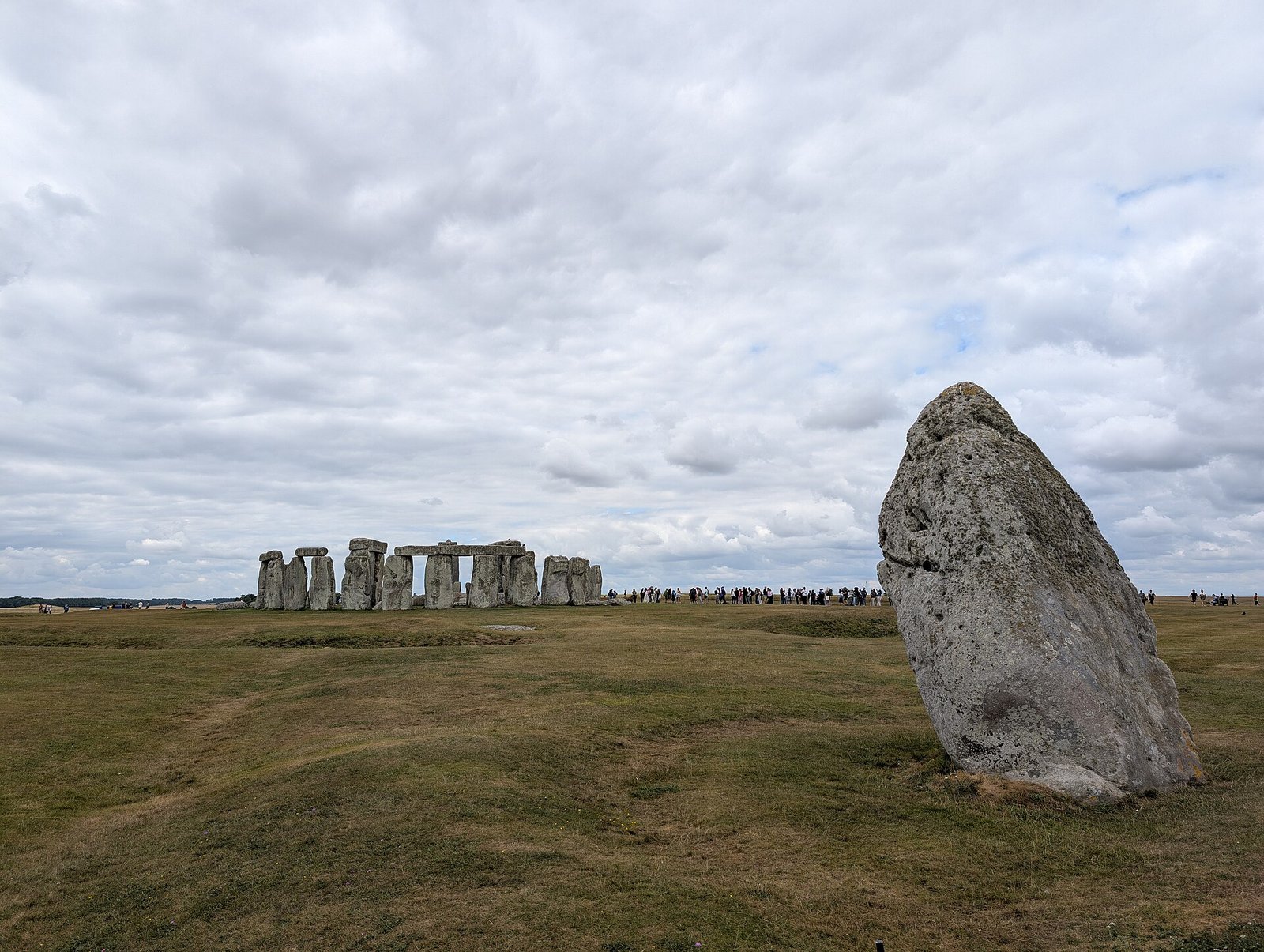 A Pedra do Calcanhar em primeiro plano diante do círculo de pedras de Stonehenge, sob céu com nuvens.