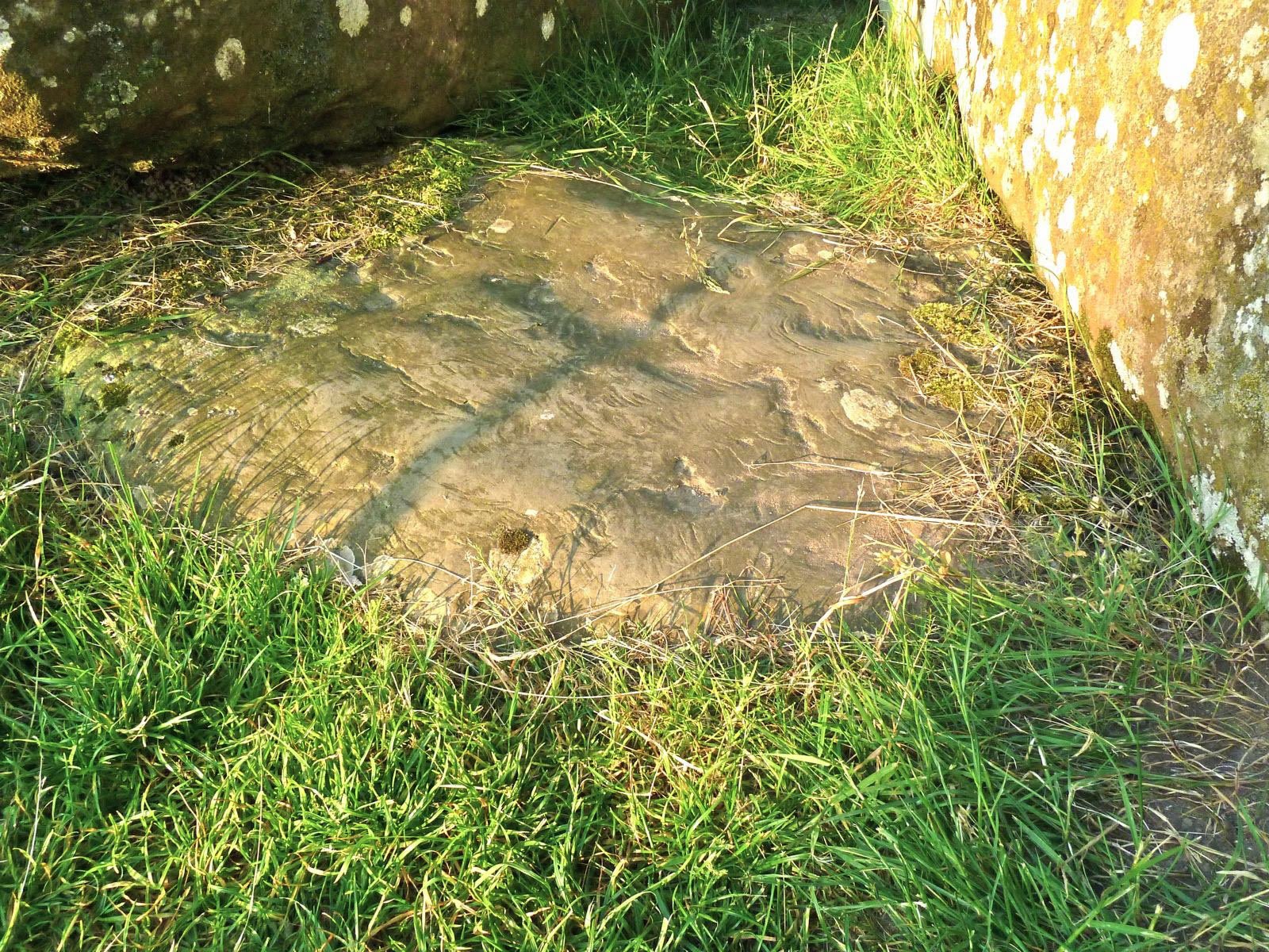 Superfície da Pedra do Altar junto ao chão, cercada de grama e parcialmente encoberta por outras pedras de Stonehenge.