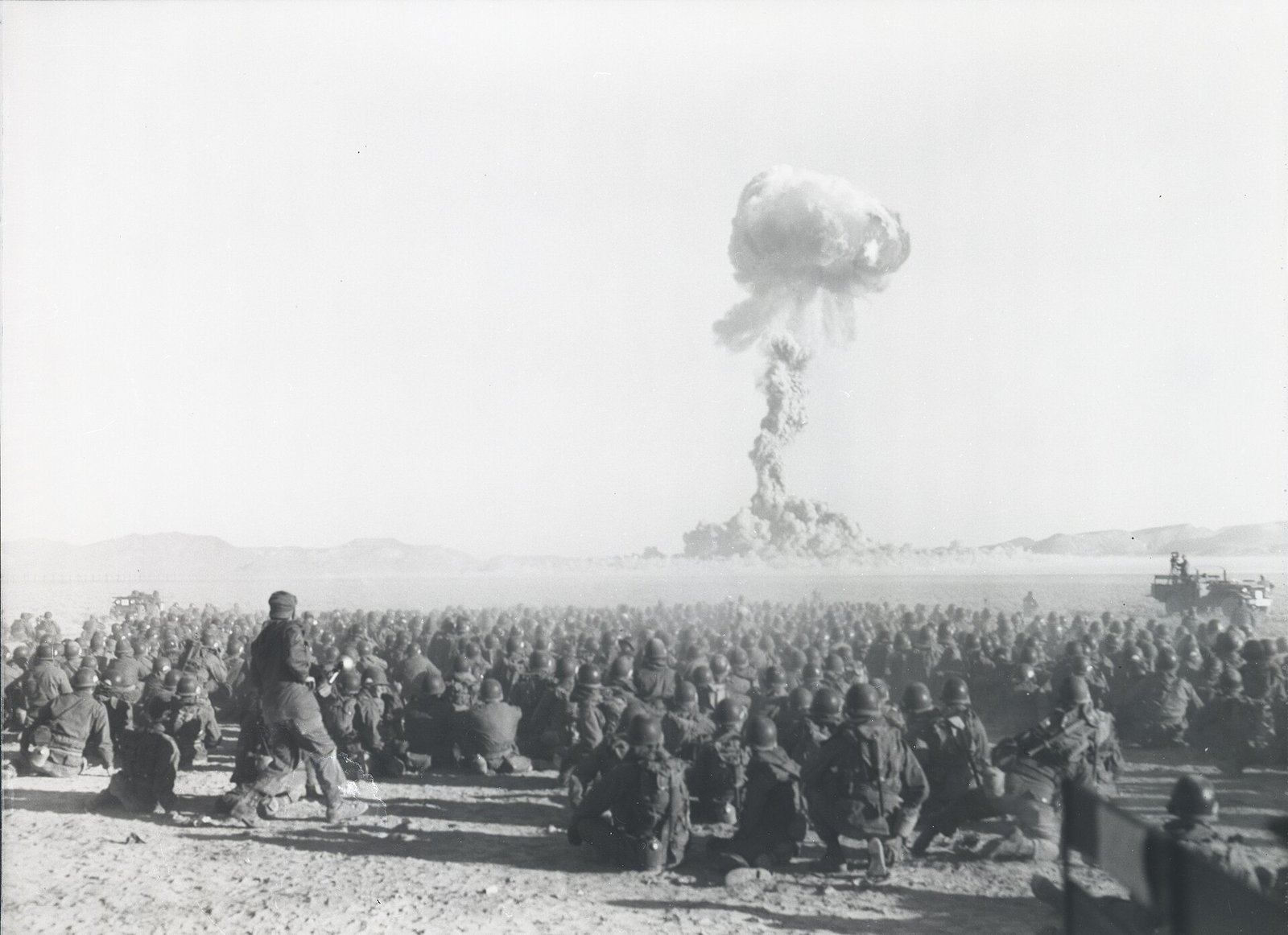 Militares observam, do deserto, a nuvem de uma explosão nuclear ao fundo.