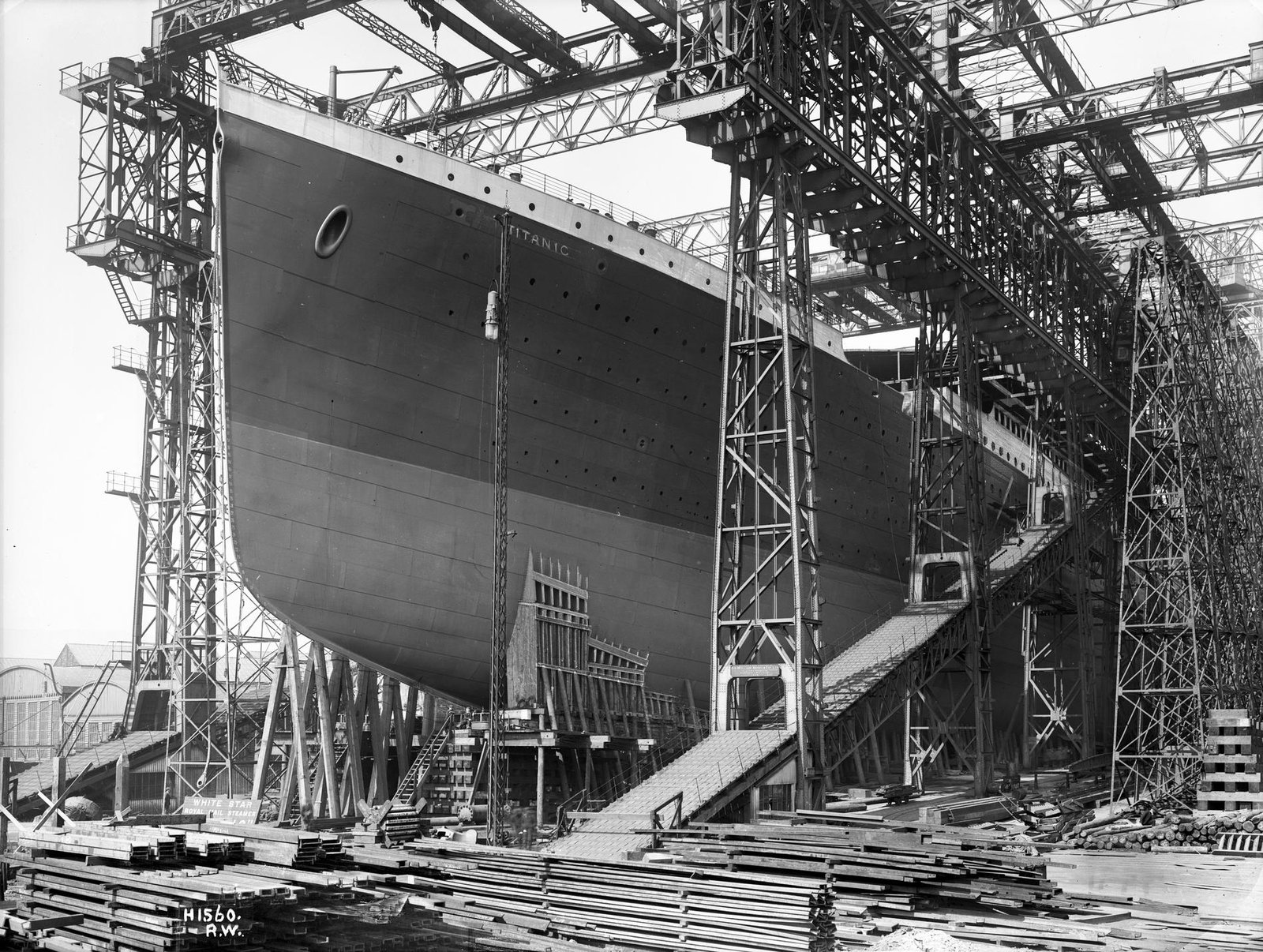 Proa do Titanic entre estruturas do estaleiro em Belfast, com trabalhadores pequenos diante do casco.
