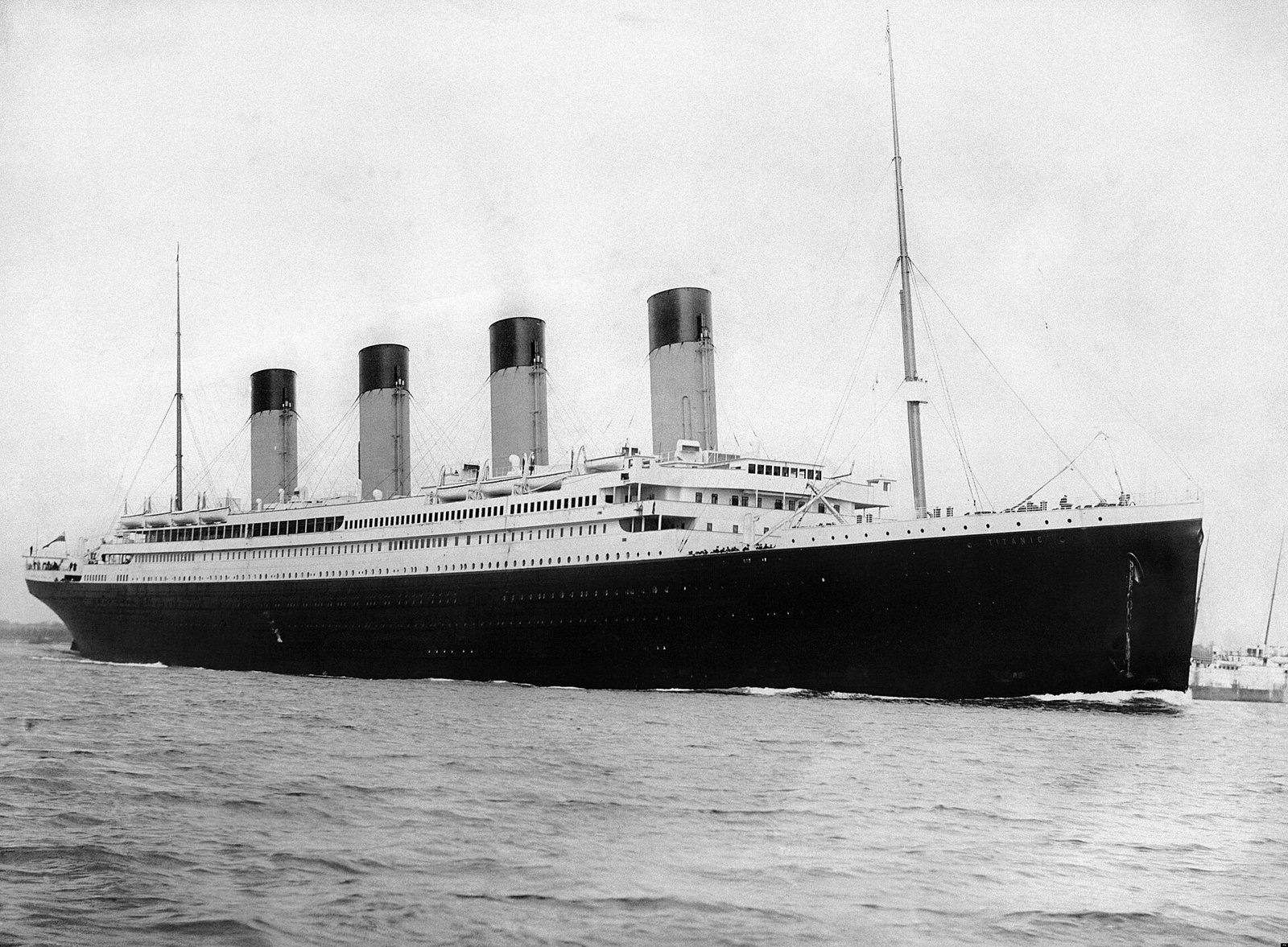 Titanic fotografado de lado, com quatro chaminés e rebocadores próximos ao casco.