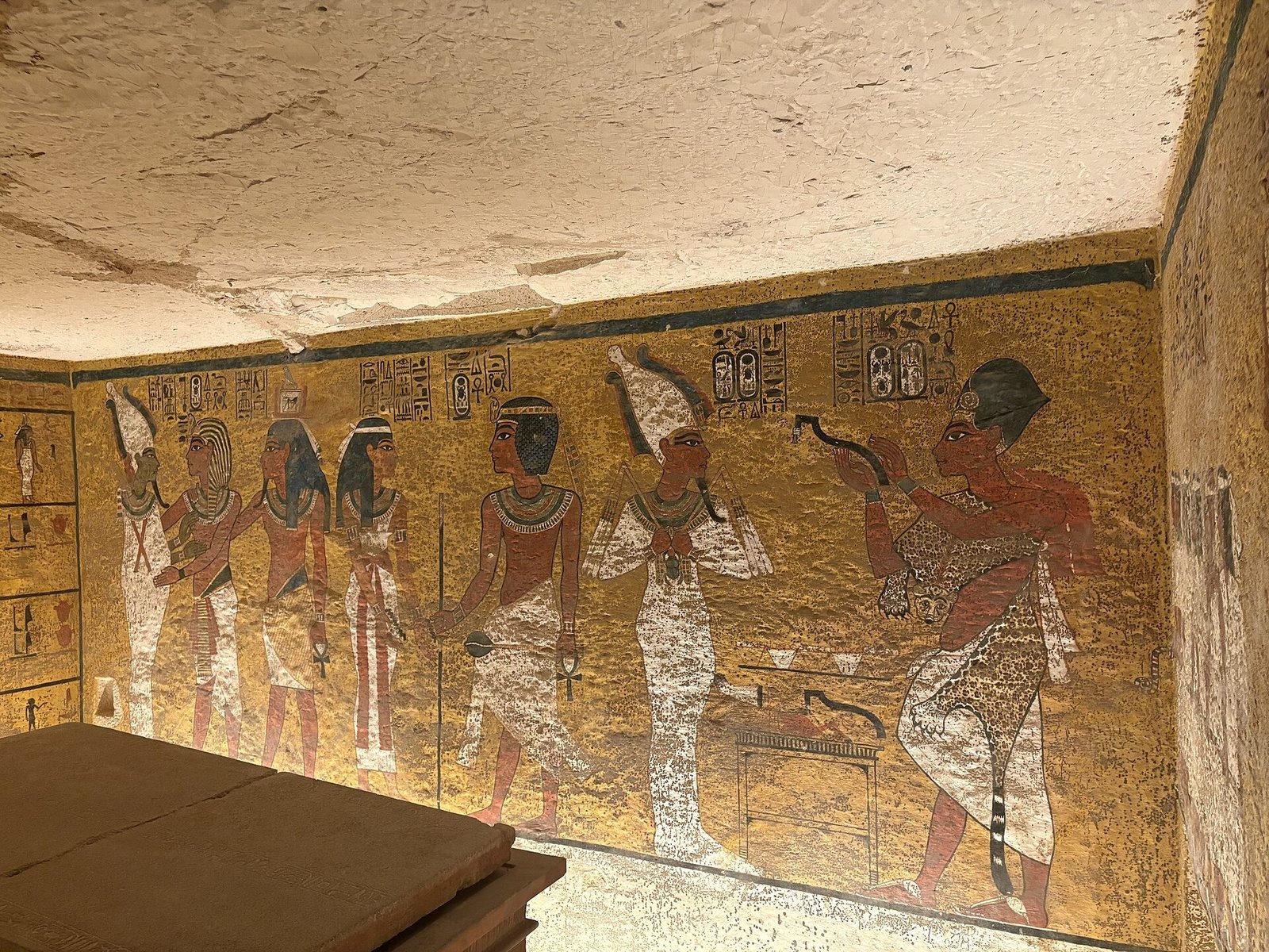 Parede pintada da câmara funerária de Tutancâmon, com figuras humanas e divinas sobre fundo amarelo e parte do sarcófago em primeiro plano.