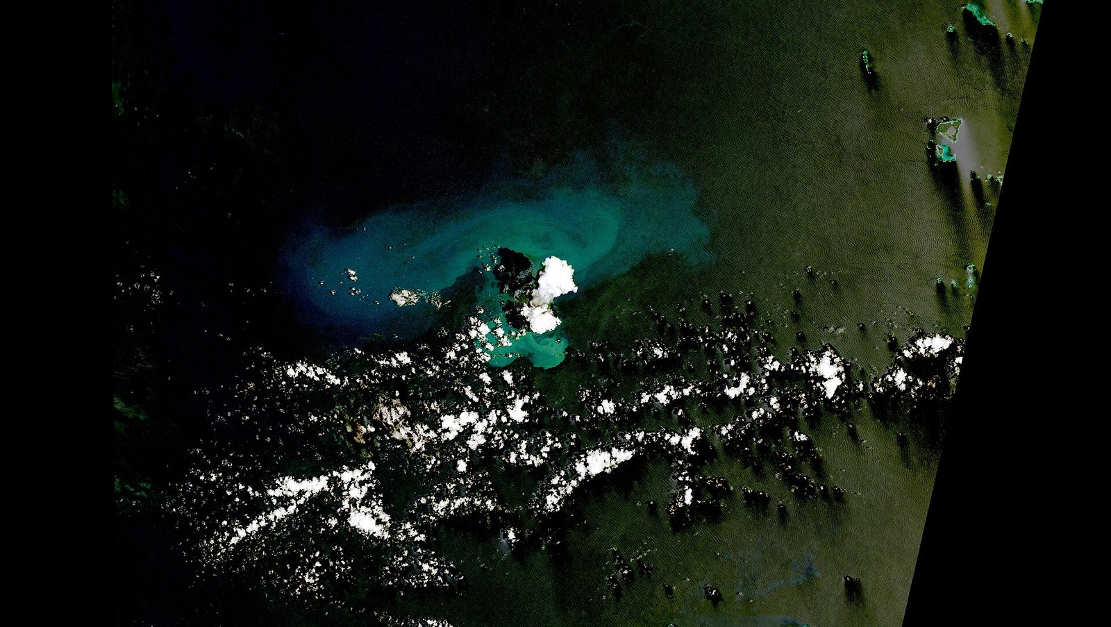 Uma pluma branca sobe de pequenas ilhas vulcânicas cercadas por água turquesa, com nuvens espalhadas sobre o oceano escuro.