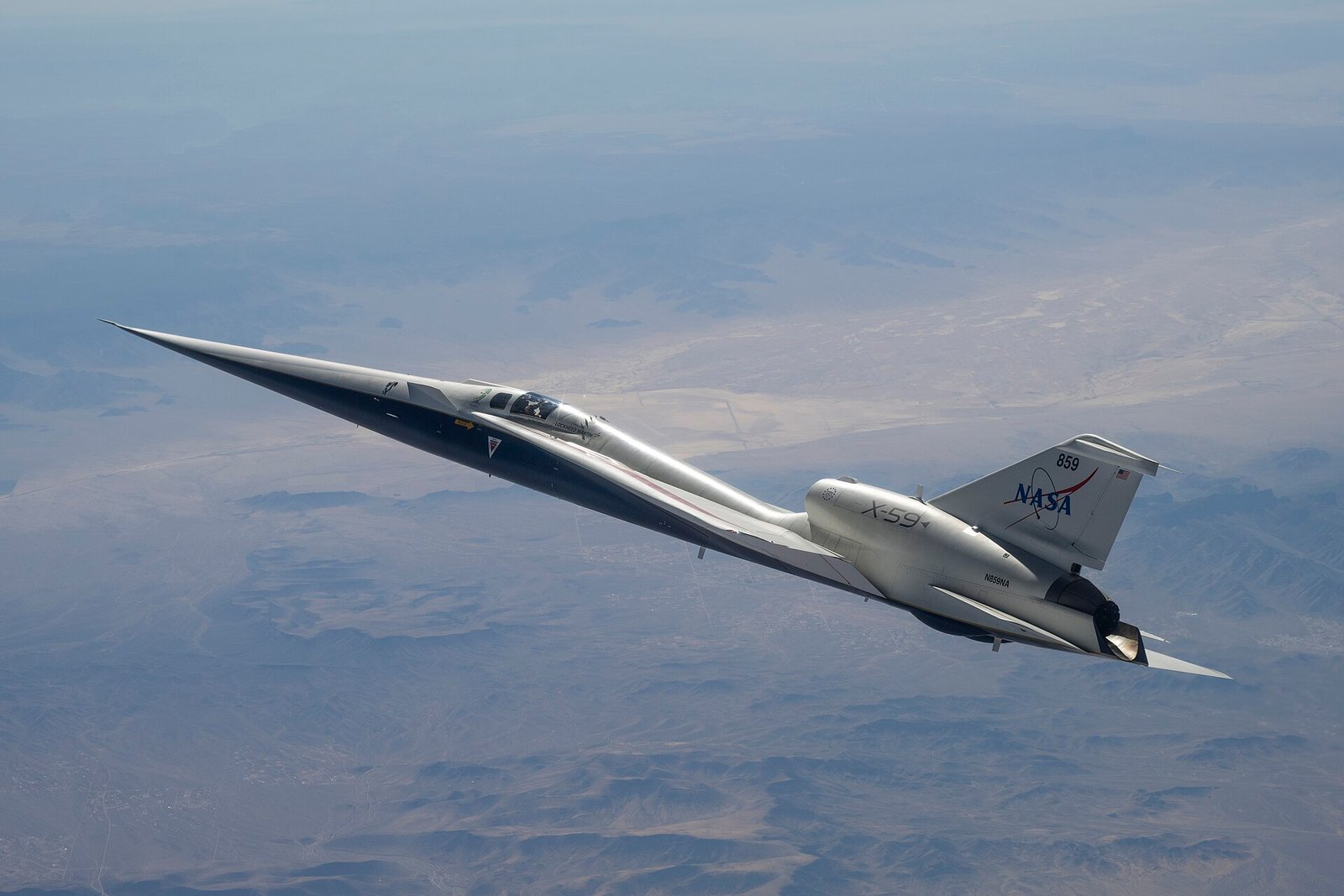 O X-59 branco e azul voa acima do deserto, com nariz muito longo, asas pequenas e motor instalado sobre a fuselagem.