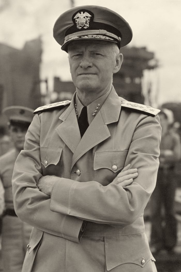 Um oficial de quepe e uniforme claro, de braços cruzados, olha sério para a câmera.