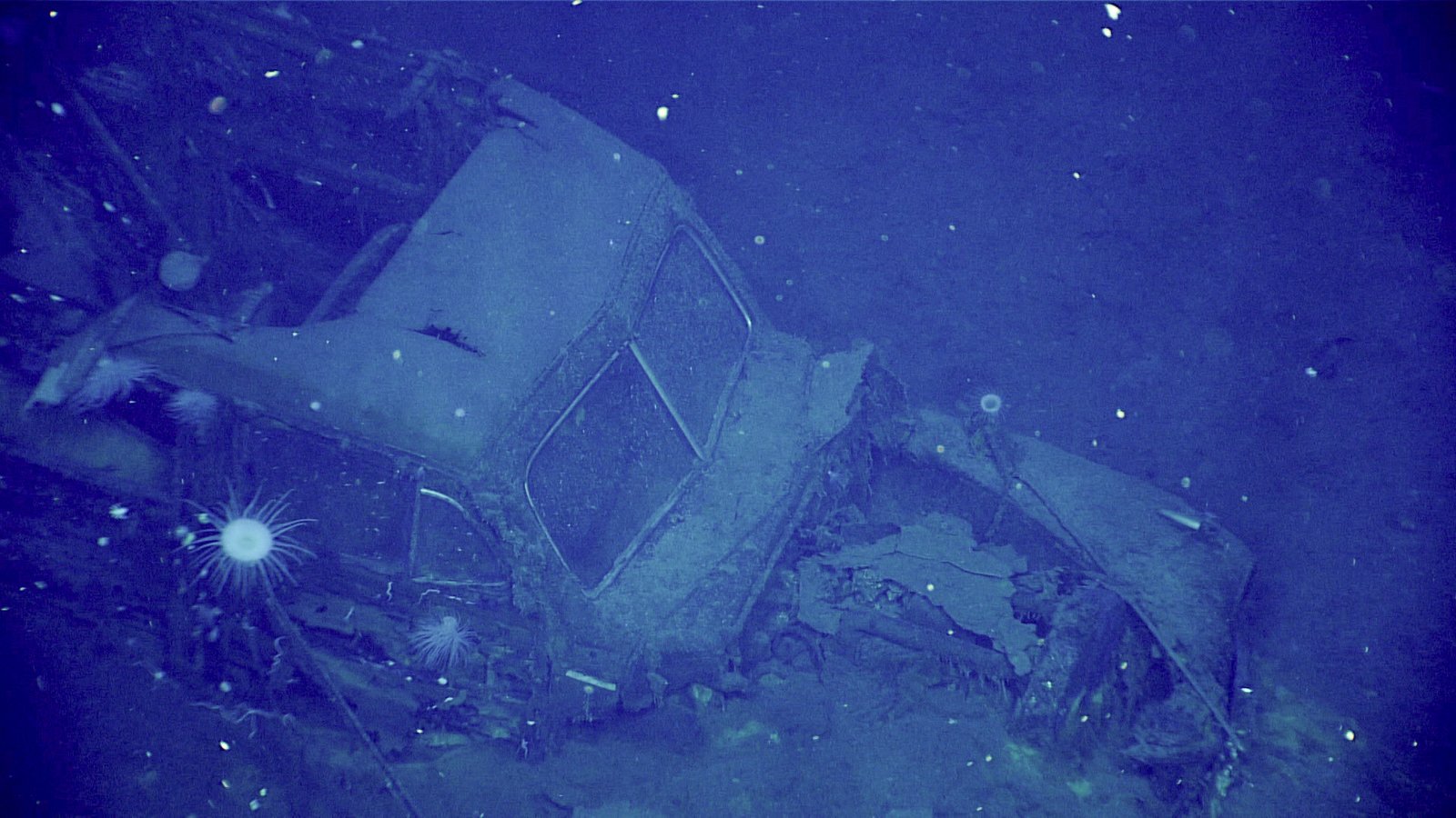 Carro deteriorado no hangar submerso do Yorktown, com para-brisa dividido e partes da carroceria iluminados pelo veículo submarino.