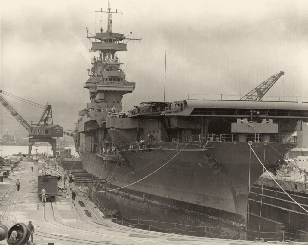 Um grande navio de guerra dentro de um dique seco, visto da proa, com guindastes, cabos e operários pequenos no cais.